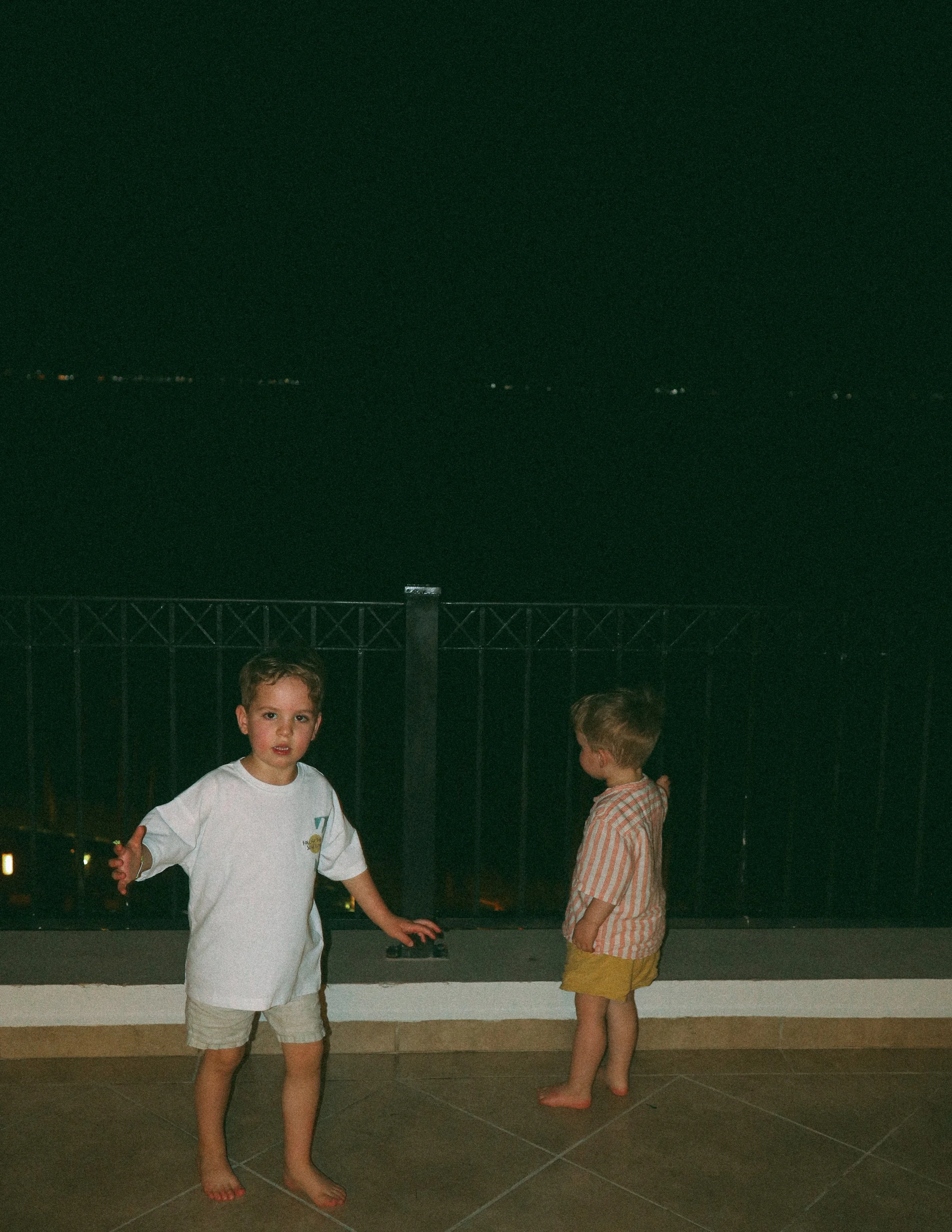 Two children standing on a tiled balcony at night, with a dark landscape in the background. The child on the left is looking forward wearing a white shirt and shorts, while the child on the right is turned to the side wearing a striped shirt and yellow shorts.