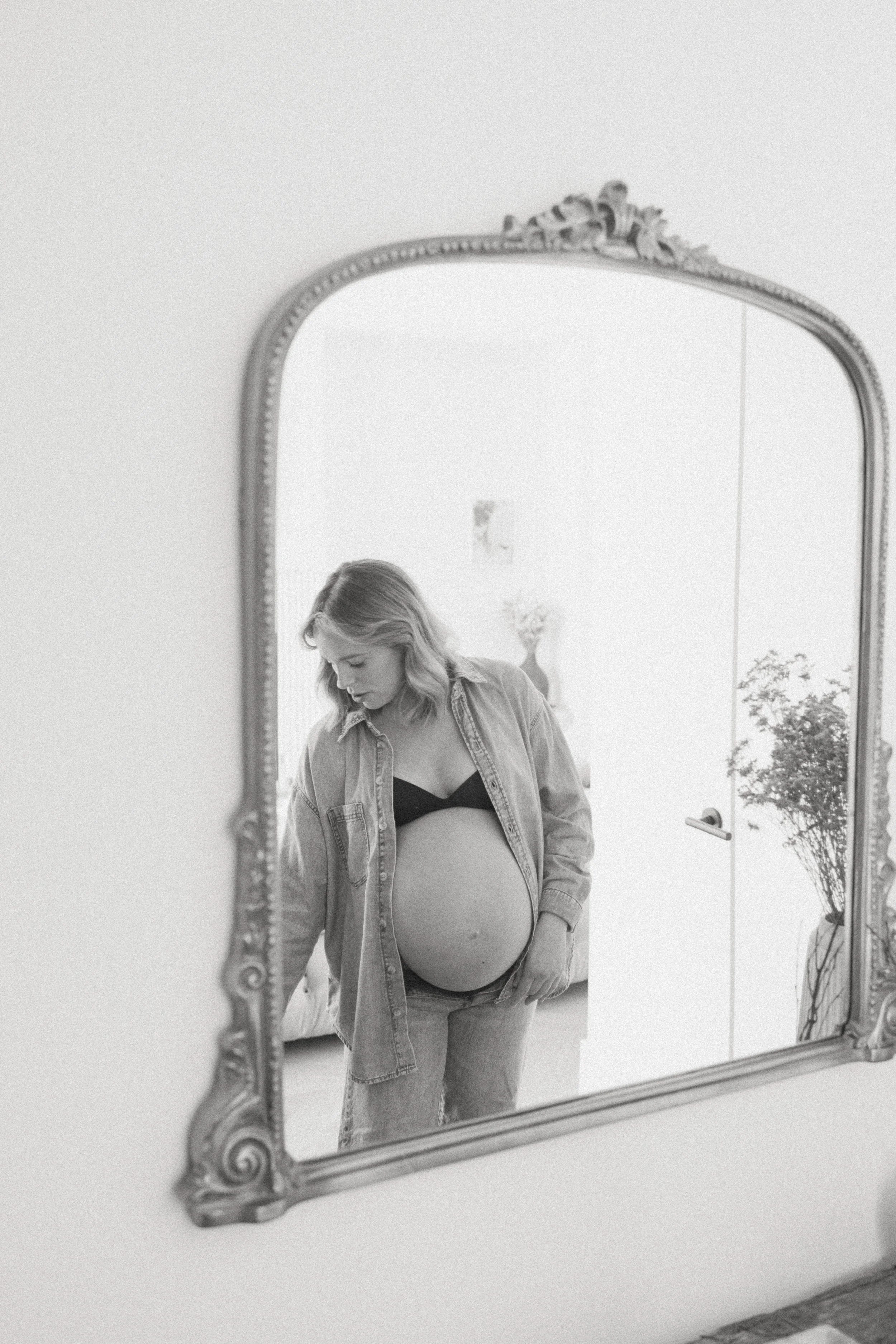 Black and white reflection of a pregnant woman in a mirror, wearing a denim shirt and black bra, with a floral vase in the background.