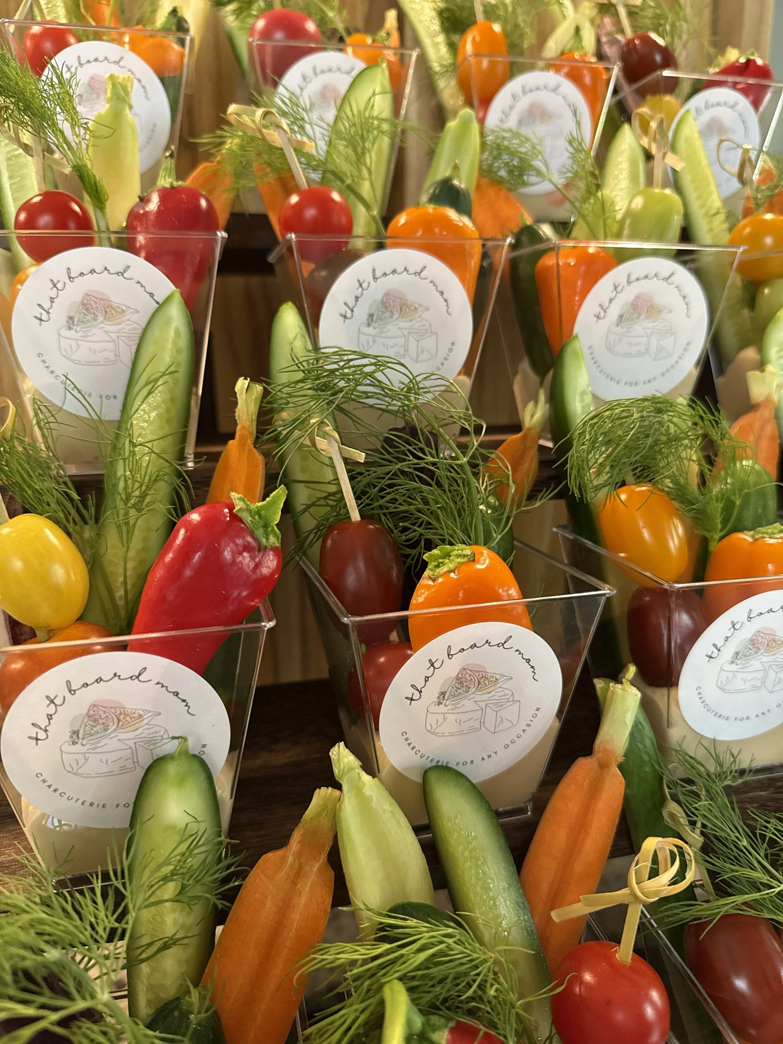 Assorted fresh vegetables, including cherry tomatoes, cucumbers, carrots, and yellow and orange bell peppers, arranged in small clear containers with a label reading "thet board mom".