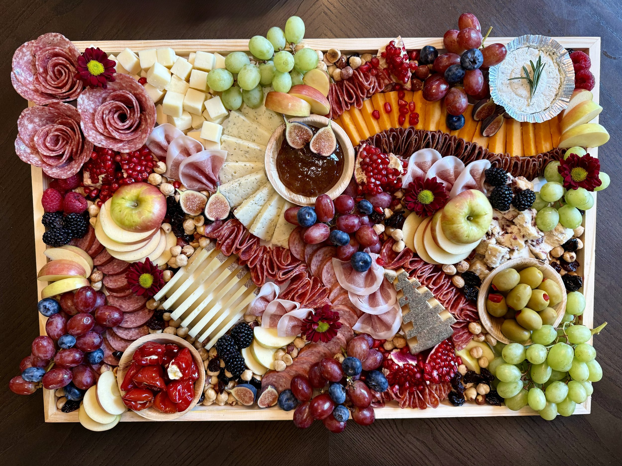 A large wooden platter with a variety of cheeses, cured meats, fruits, nuts, and garnishes arranged for a charcuterie board.