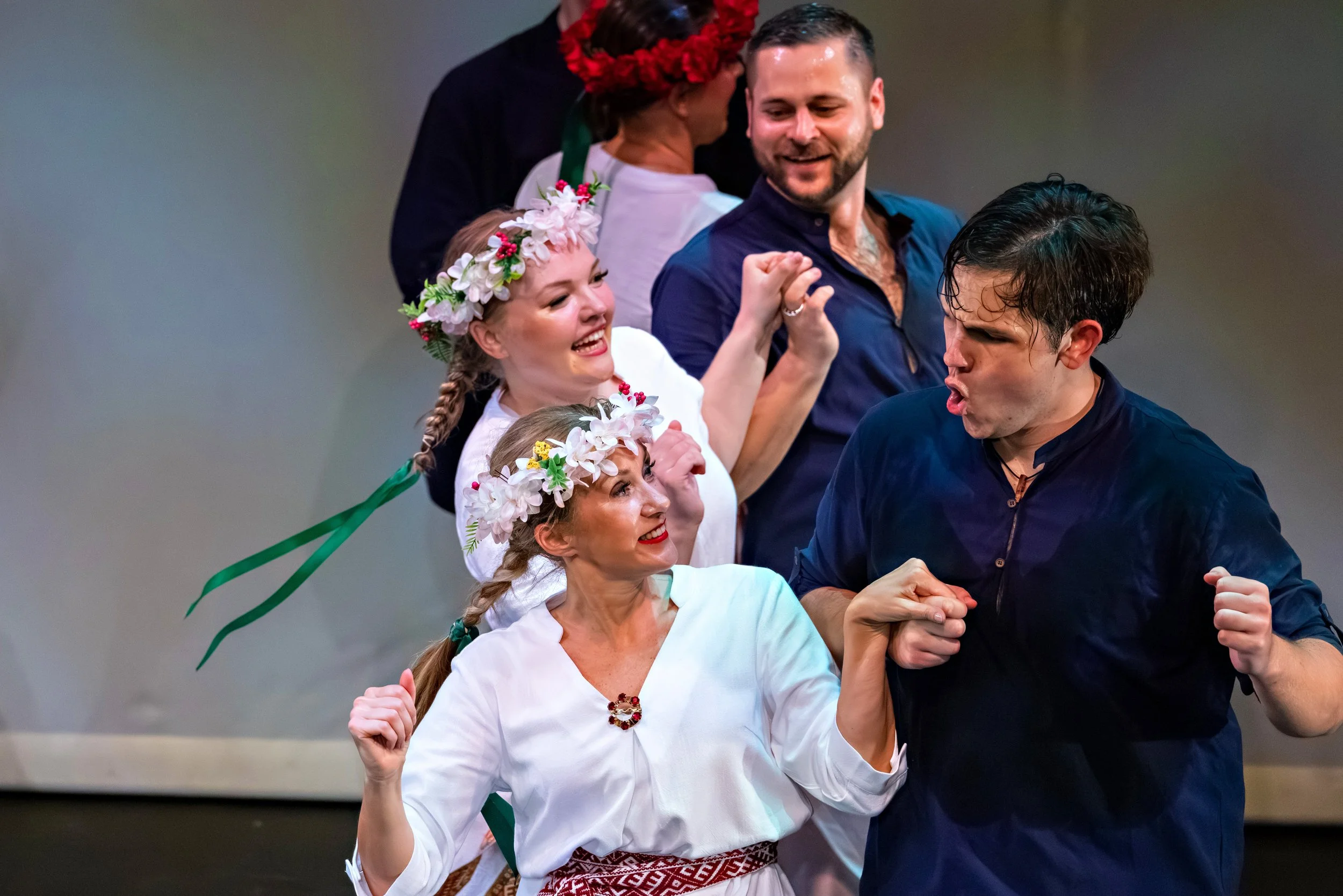 Group of people celebrating, dressed in traditional clothing; two women wearing flower crowns and smiling, a man and woman in dark clothing showing animated expressions, possibly at a cultural or festive event.