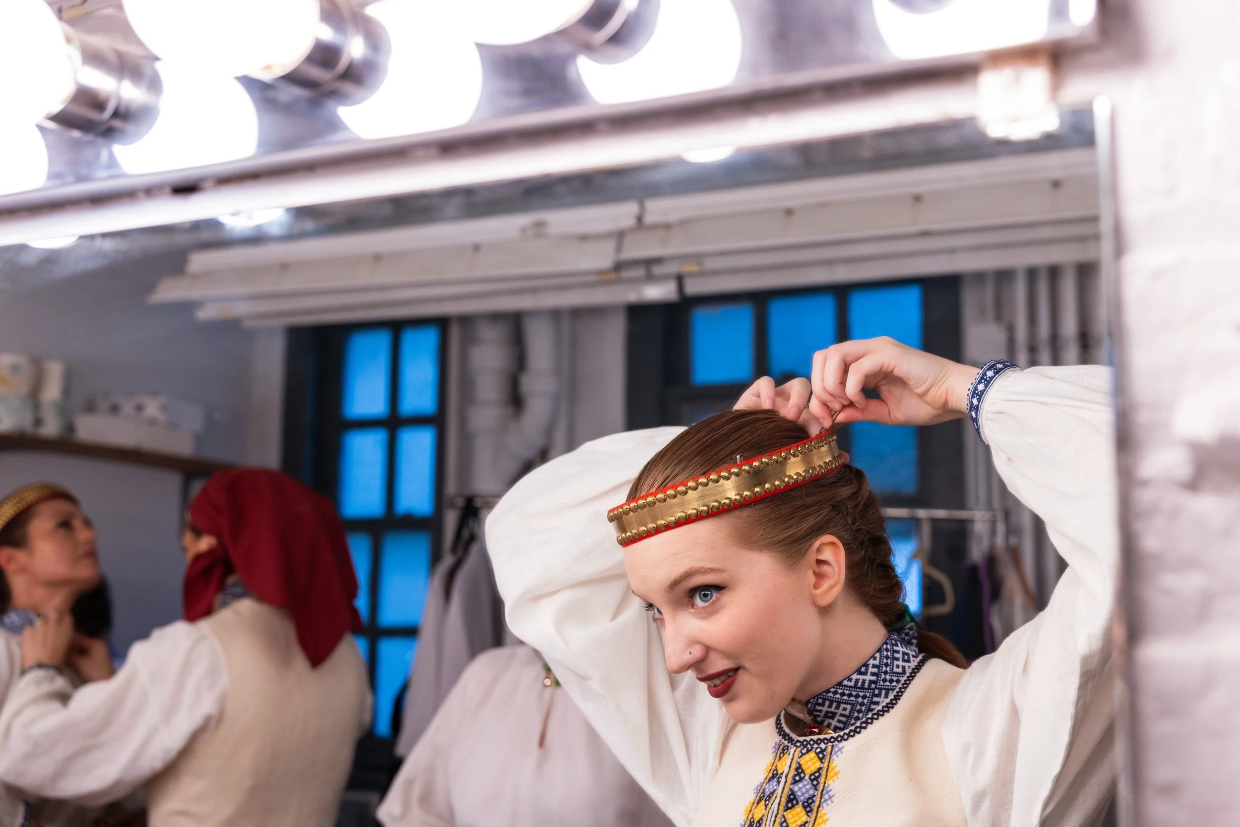 A woman in traditional Latvian folk costume adjusts a decorative headband, with other women in similar costumes in the background, preparing for a cultural performance.
