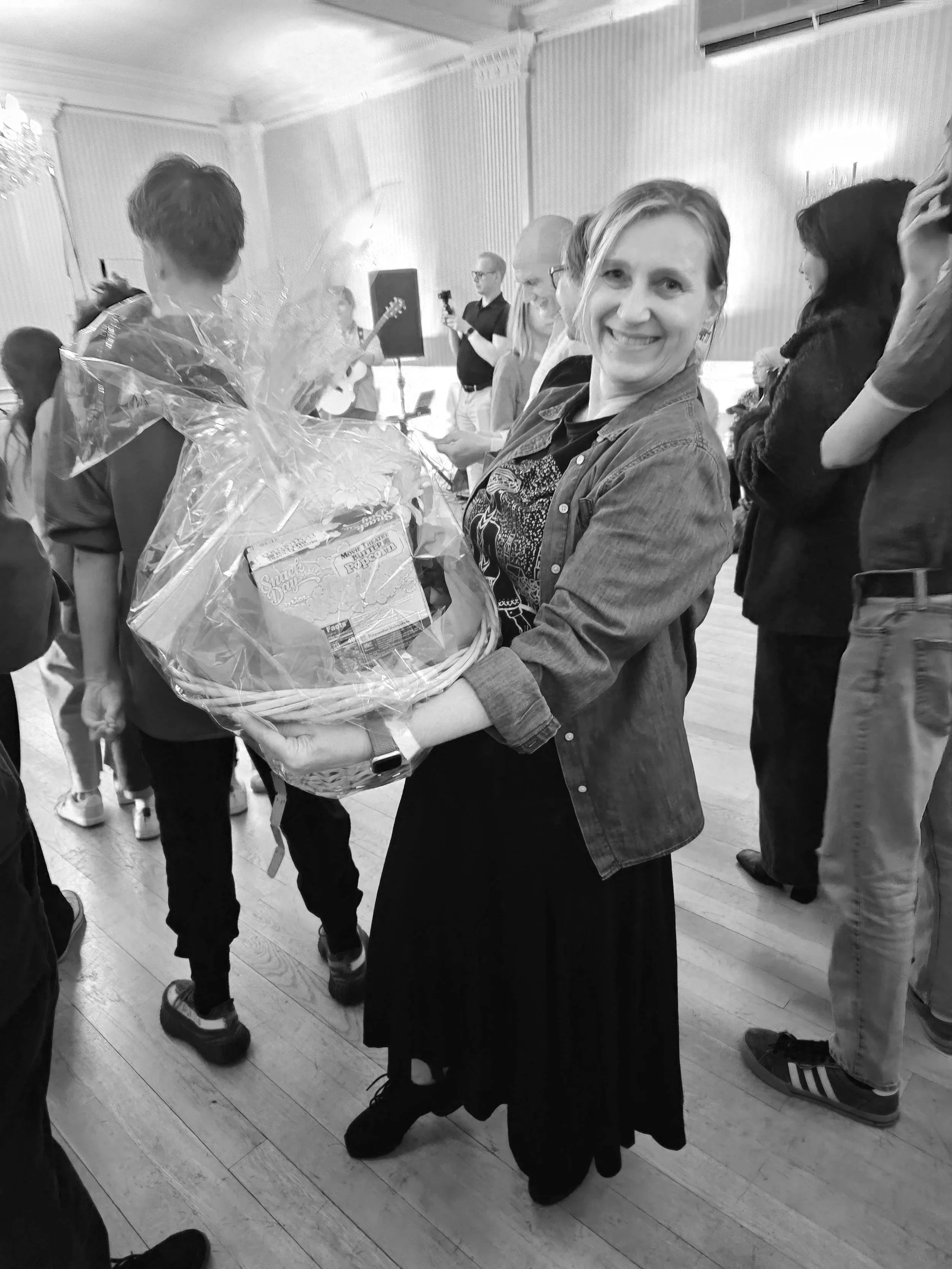 A woman smiling and holding a large gift basket wrapped in cellophane at a social gathering, with several people in the background.