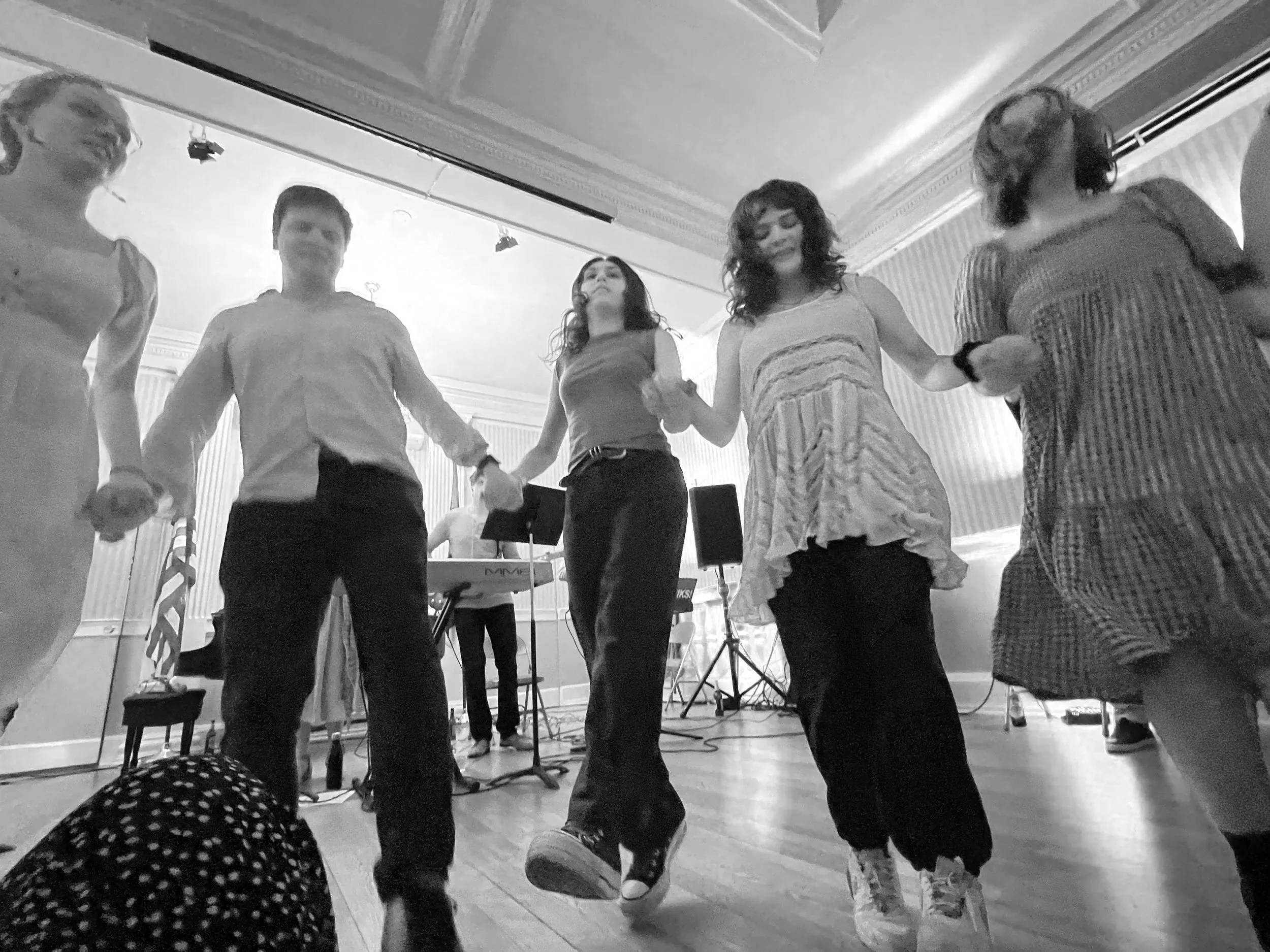 People dancing in a line at an indoor event with a band setup in the background.