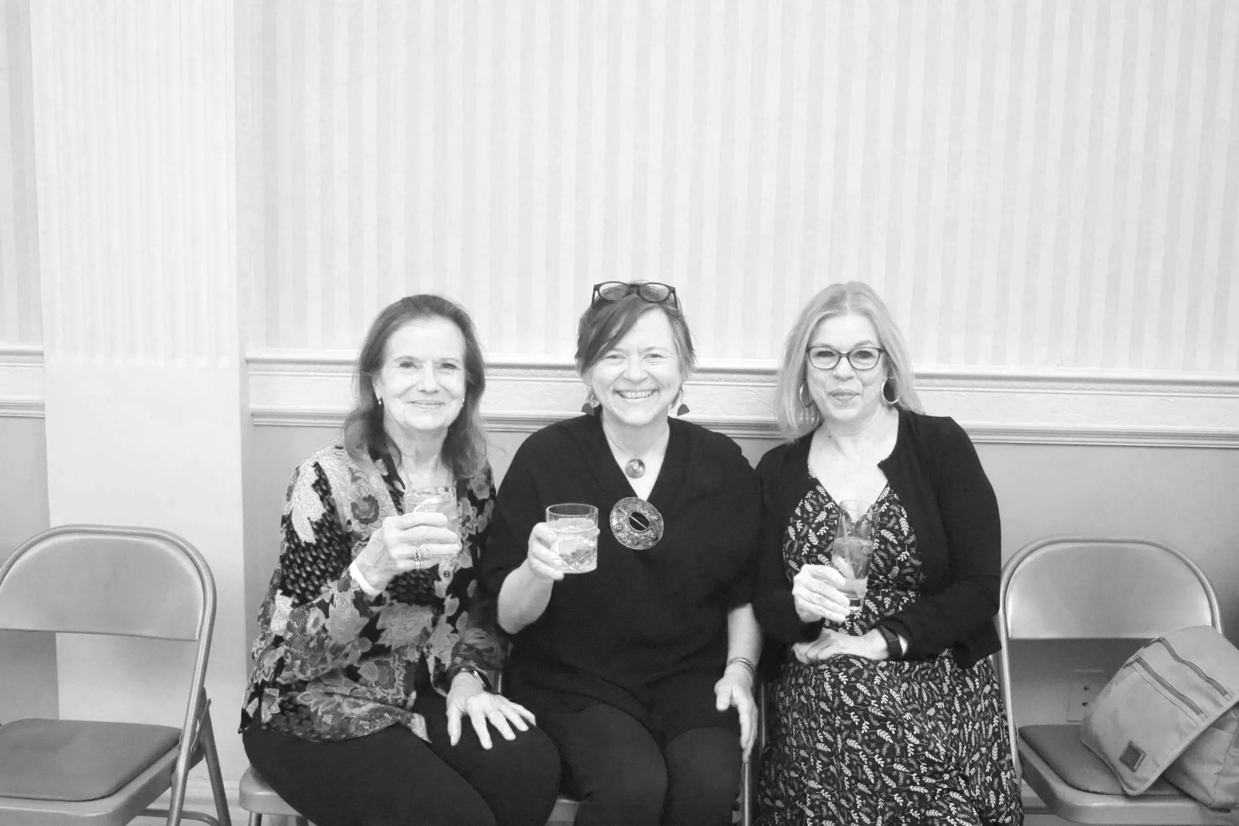 Three women sitting on chairs, holding drinks and smiling at the camera in a room with striped wallpaper.