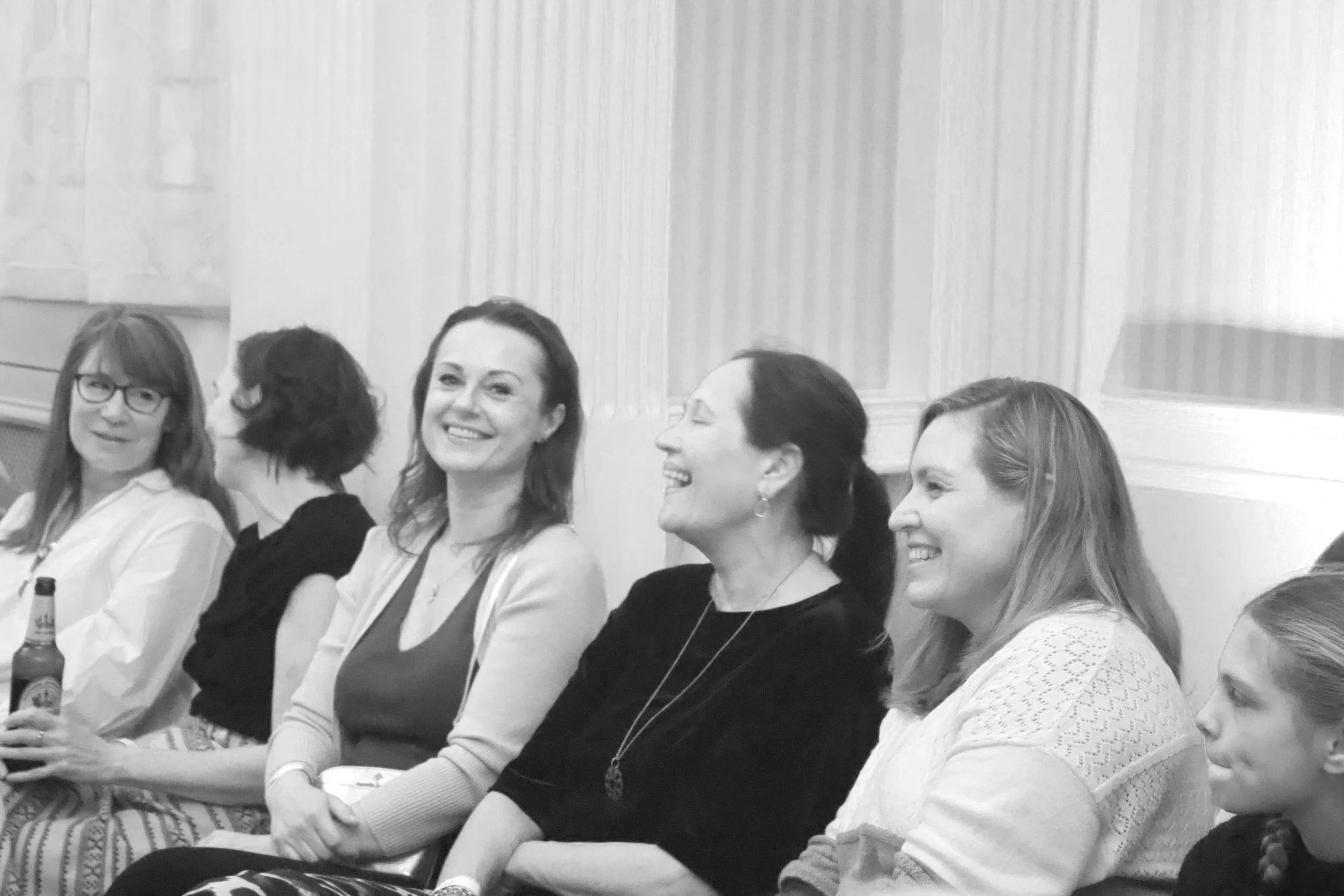 Black and white photo of women sitting in a row, smiling and laughing at a social gathering or event.