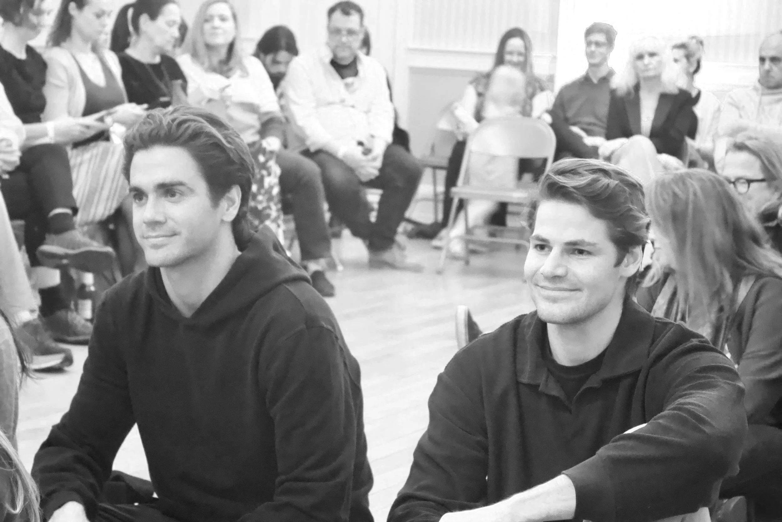 People seated on the floor and chairs in a room, listening attentively, with two young men in the foreground.