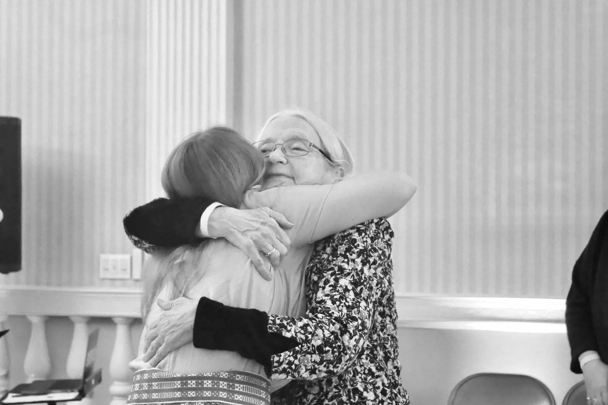 Three women sharing an embrace at an indoor event, with a wall with vertical stripes in the background.