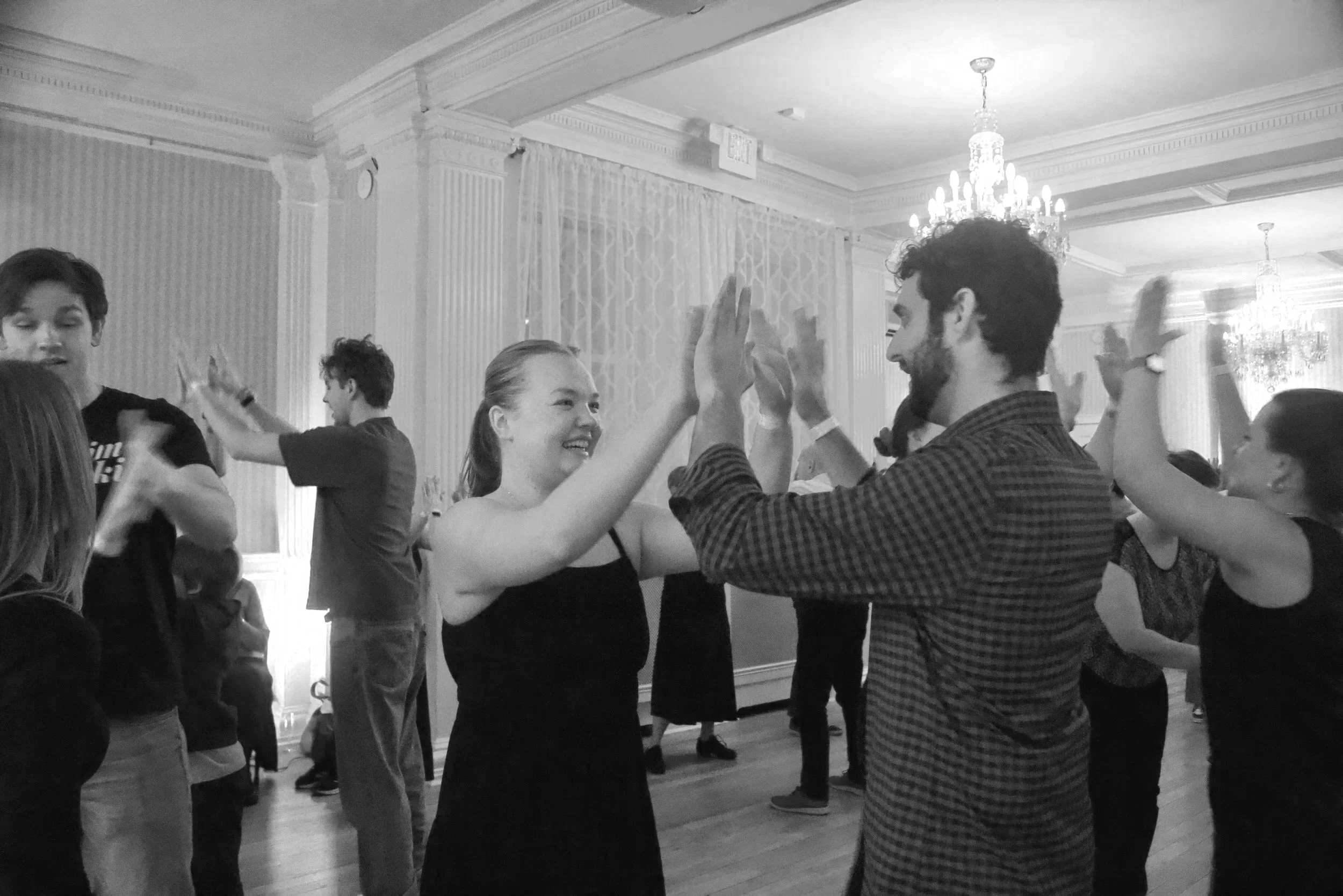 People dancing and smiling at an indoor party with chandeliers and elegant curtains.