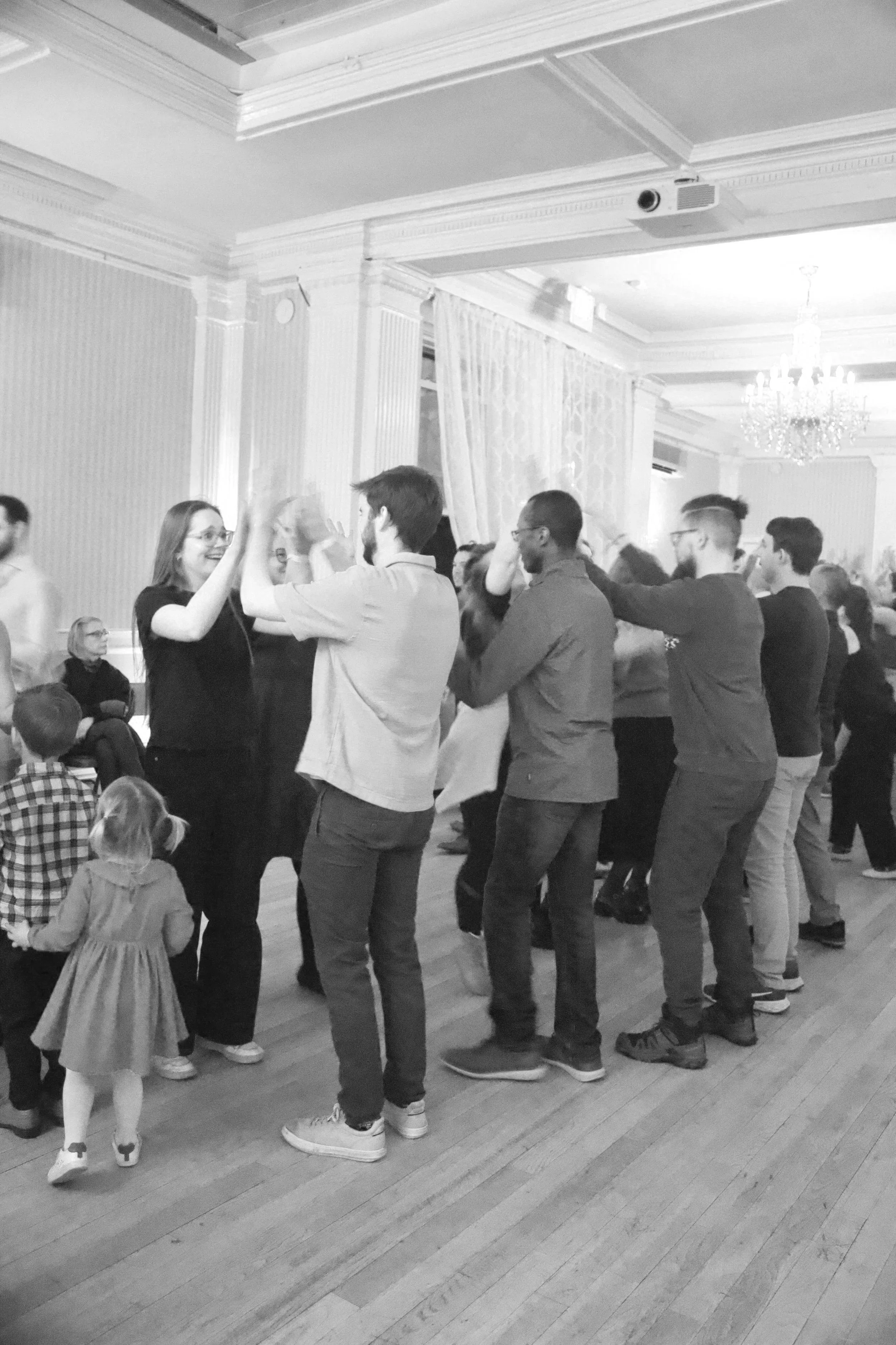 People are dancing in a line at a celebration, with children watching nearby in a large, ornate room with chandeliers and detailed crown molding.