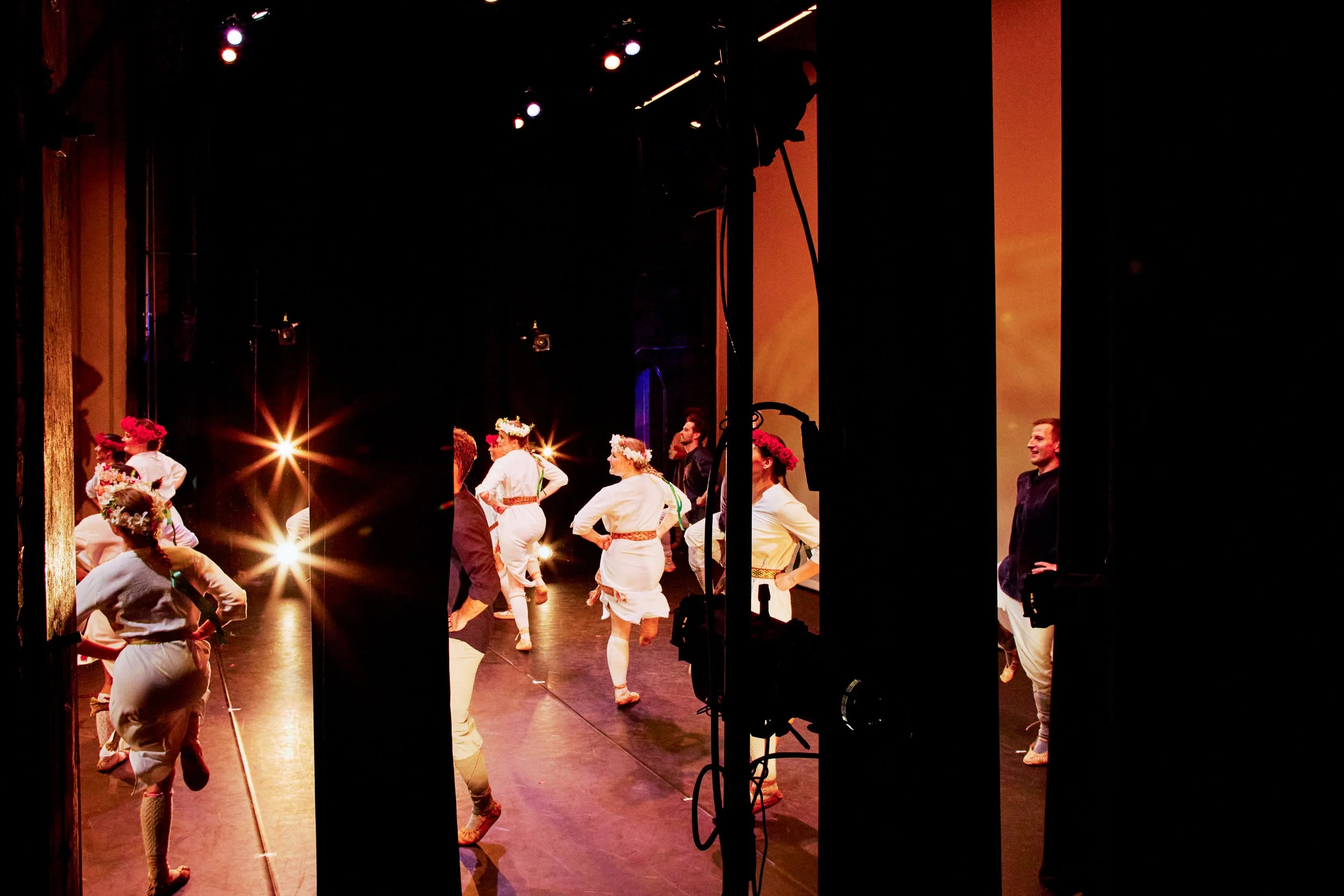 People on stage preparing for a dance performance, with some wearing floral crowns and traditional costumes, backstage view with lighting equipment.