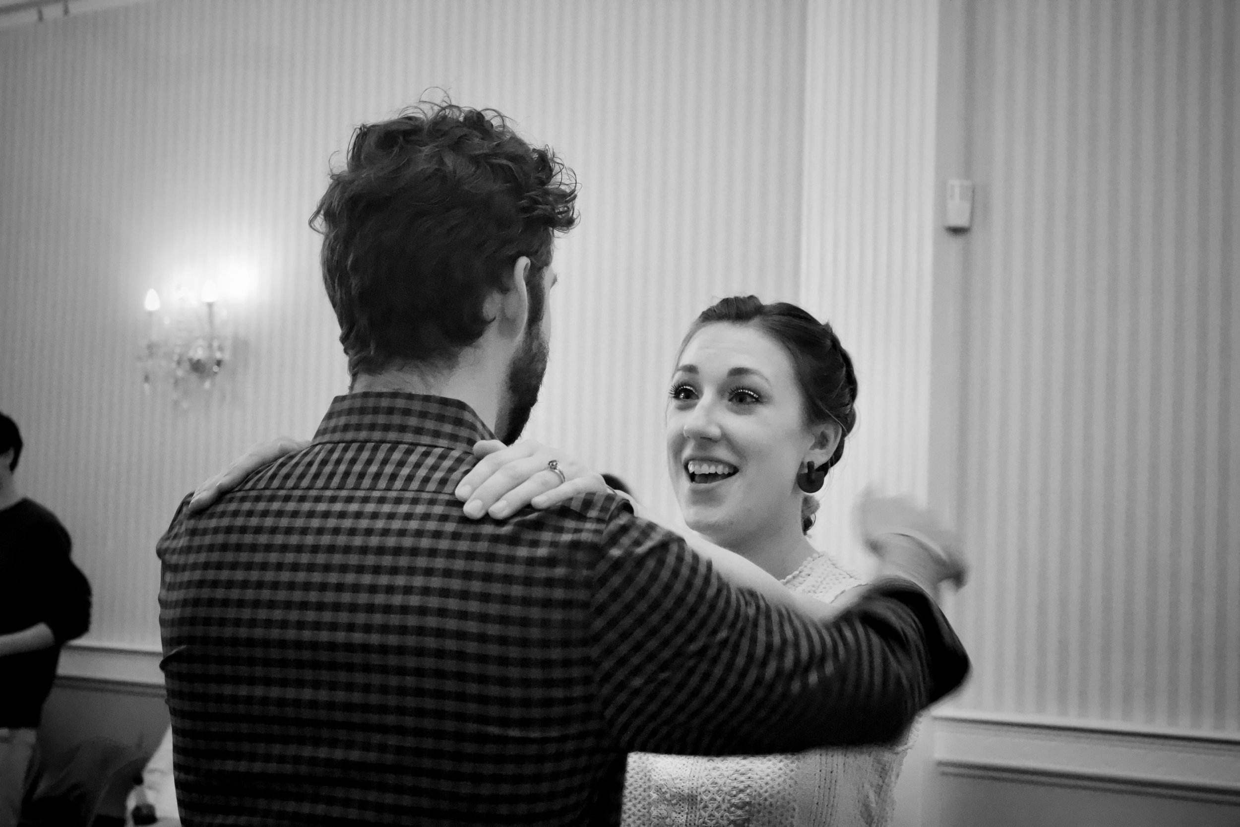 A man and woman dancing together at an indoor event, smiling and looking at each other, with a woman in the background and wall sconce lighting.