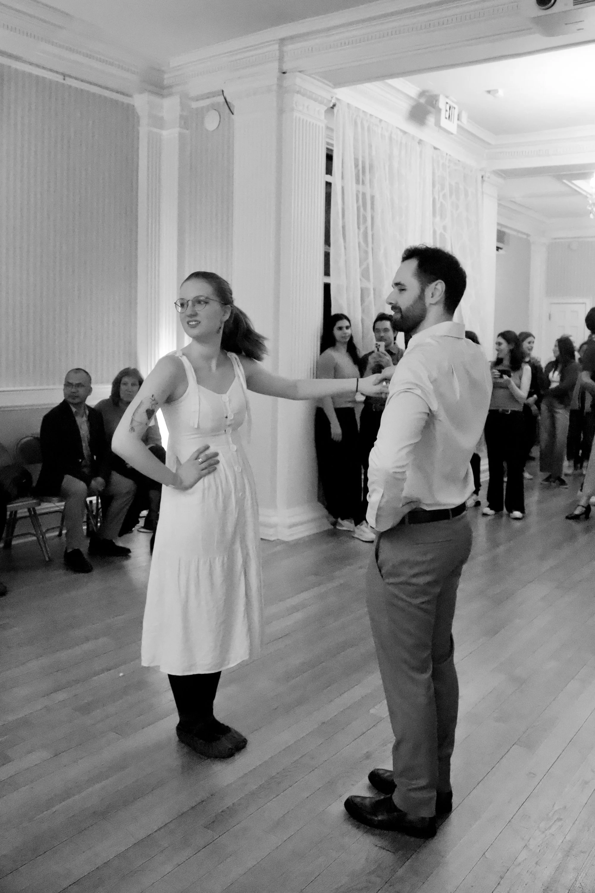 A black-and-white photo of a woman and man dancing in a room with many people watching, with some seated and some standing, and ornate decor.