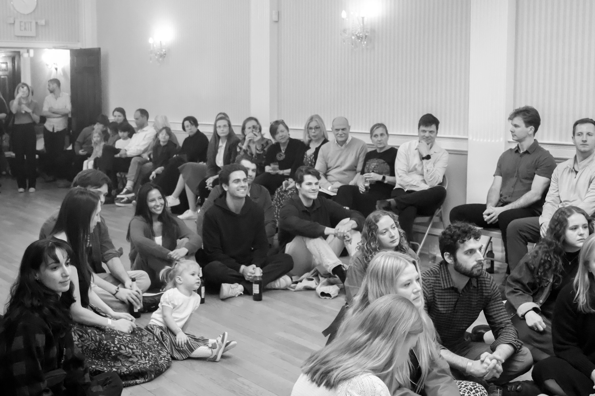 Group of people sitting and standing in a room, some watching something, including children, adults, and young adults, in a casual setting