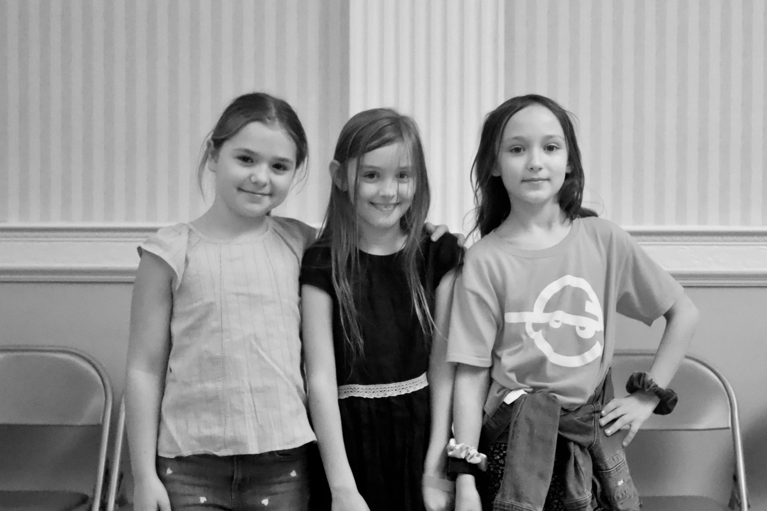 Three young girls standing together and smiling for a photo in an indoor setting with chairs behind them.