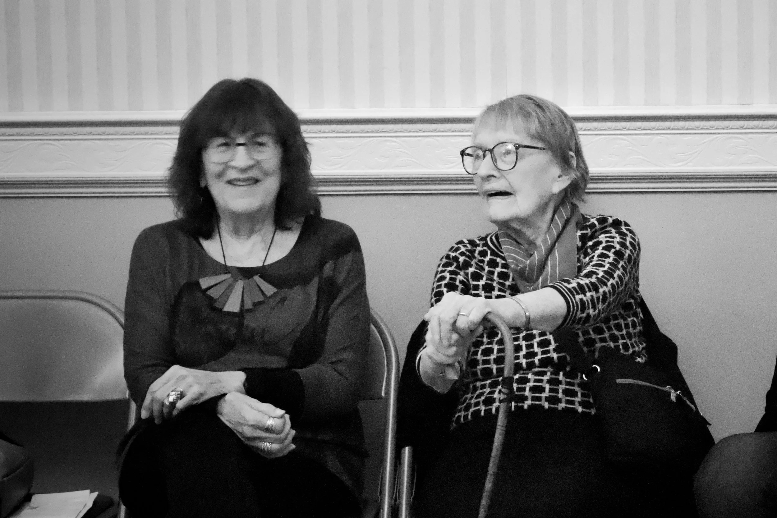 Two elderly women sitting side by side on chairs, smiling and engaging in conversation. One woman has dark hair and glasses, the other has light hair, glasses, and is holding a cane.