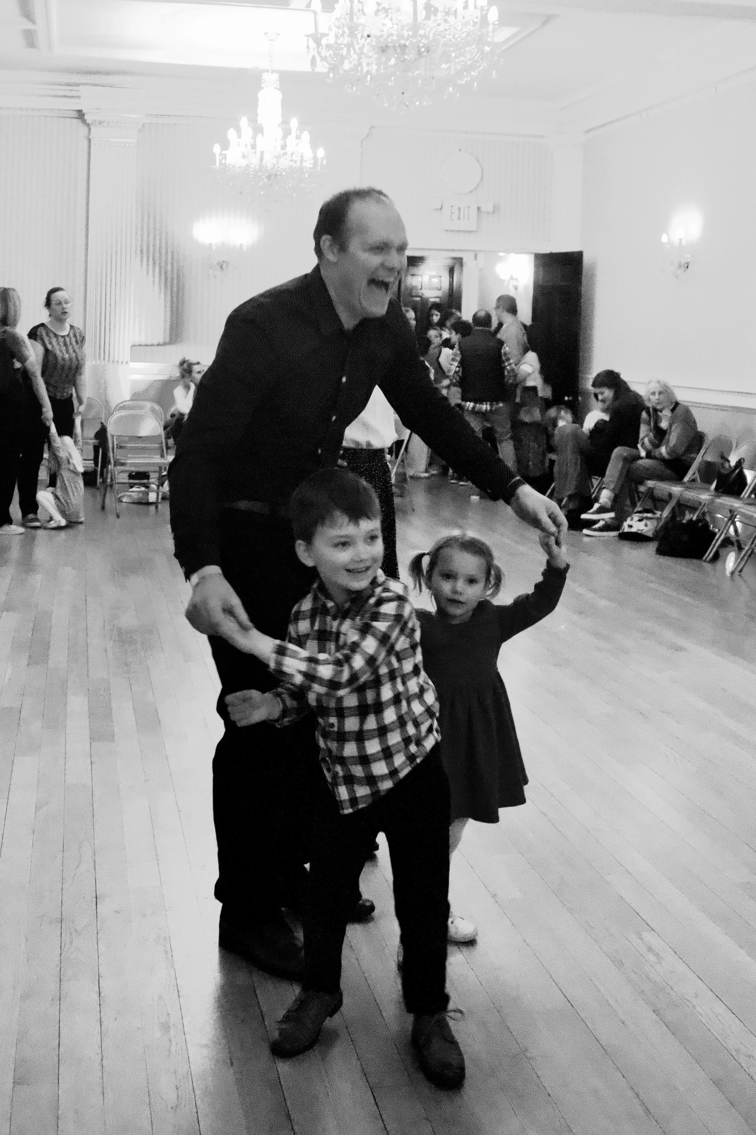 A man and two young girls dancing and holding hands in a large room with a wooden floor, chandeliers, and several people sitting or standing in the background.