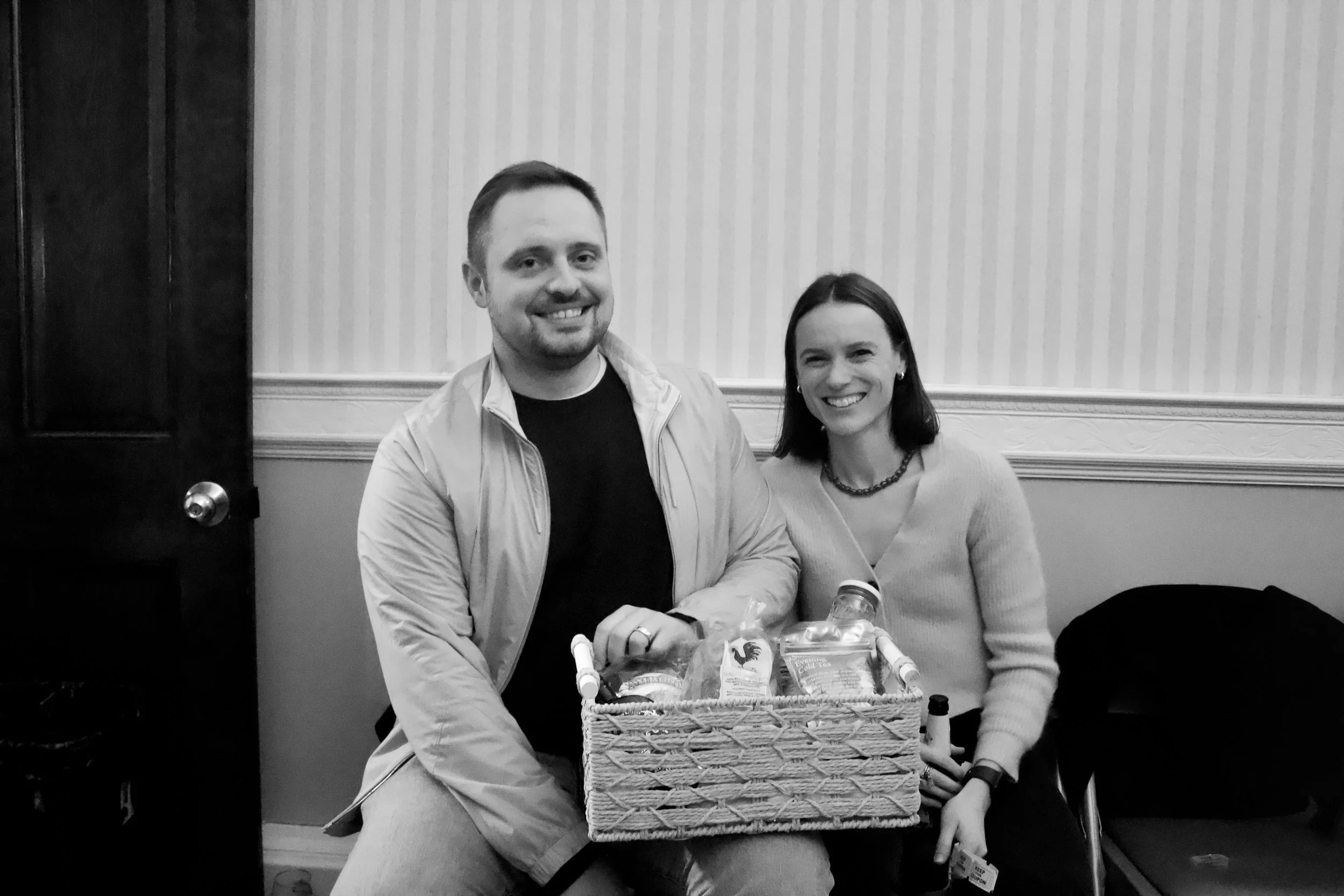 A black and white photo of a smiling man and woman sitting together indoors. The man has short hair and a beard, wearing a casual jacket and t-shirt. The woman has shoulder-length hair, wearing a sweater and necklace. They are holding a basket filled