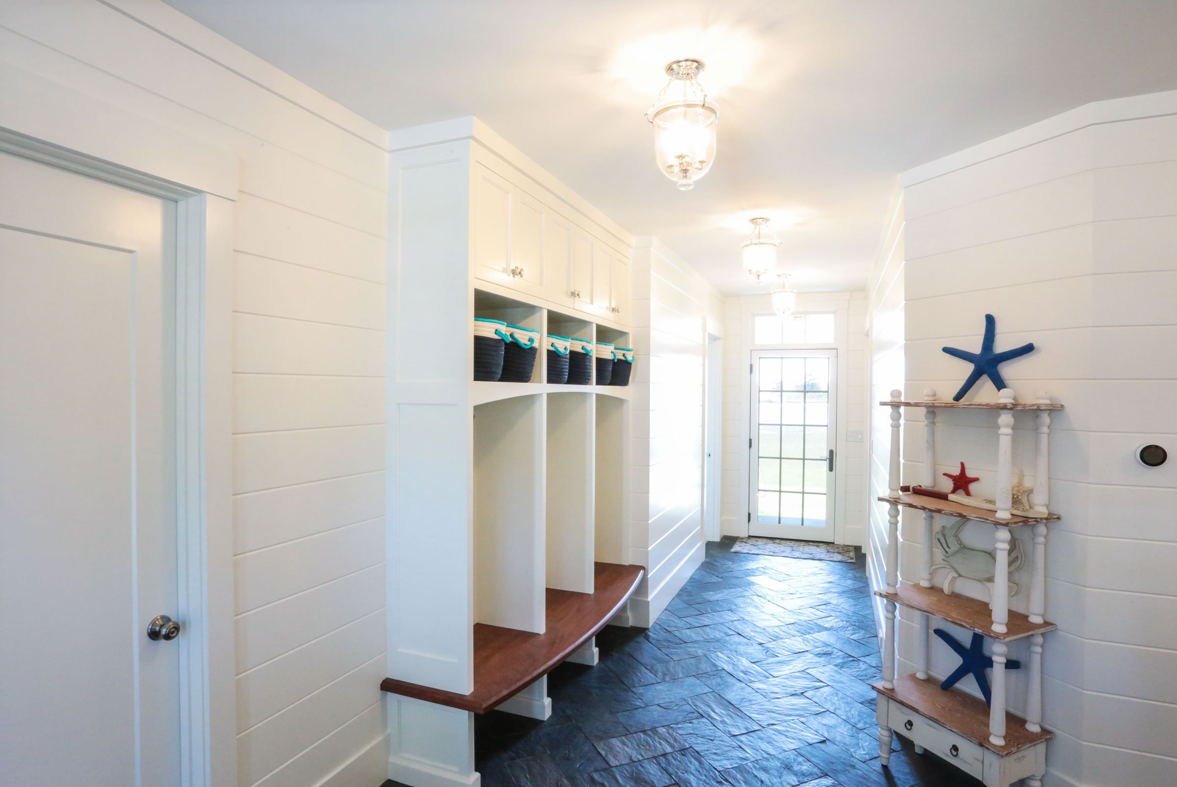 A bright and thoughtfully designed mudroom by Lindsey Architects, featuring custom built-in storage, shiplap wall detailing, and durable natural stone flooring inspired by timeless New England coastal living. Integrated bench seating, organized cubbi