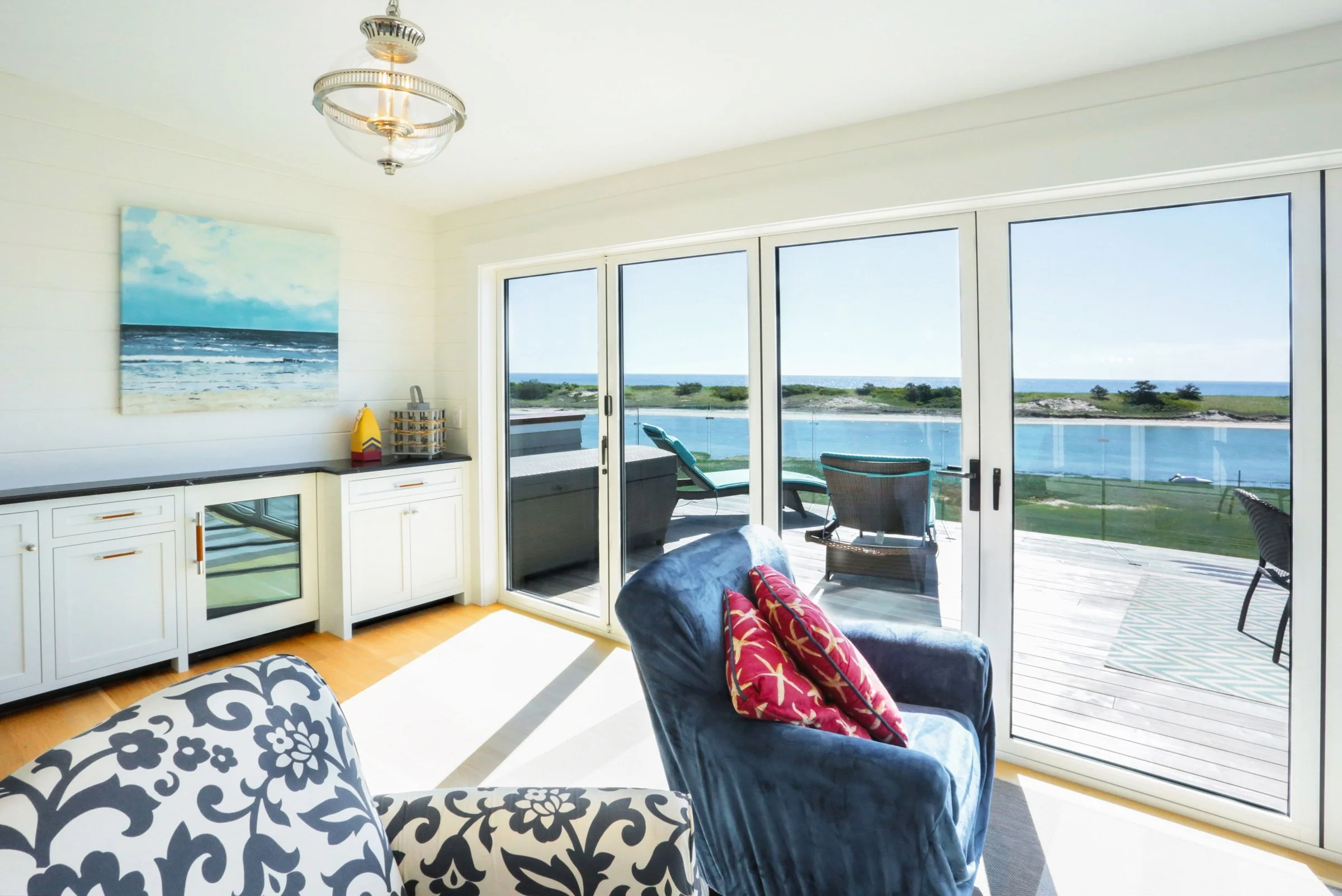 A bright and elegant waterfront sitting room designed by Lindsey Architects, featuring expansive sliding glass doors, panoramic coastal views, and seamless indoor-outdoor living inspired by the Maine and New Hampshire shoreline. Soft natural finishes