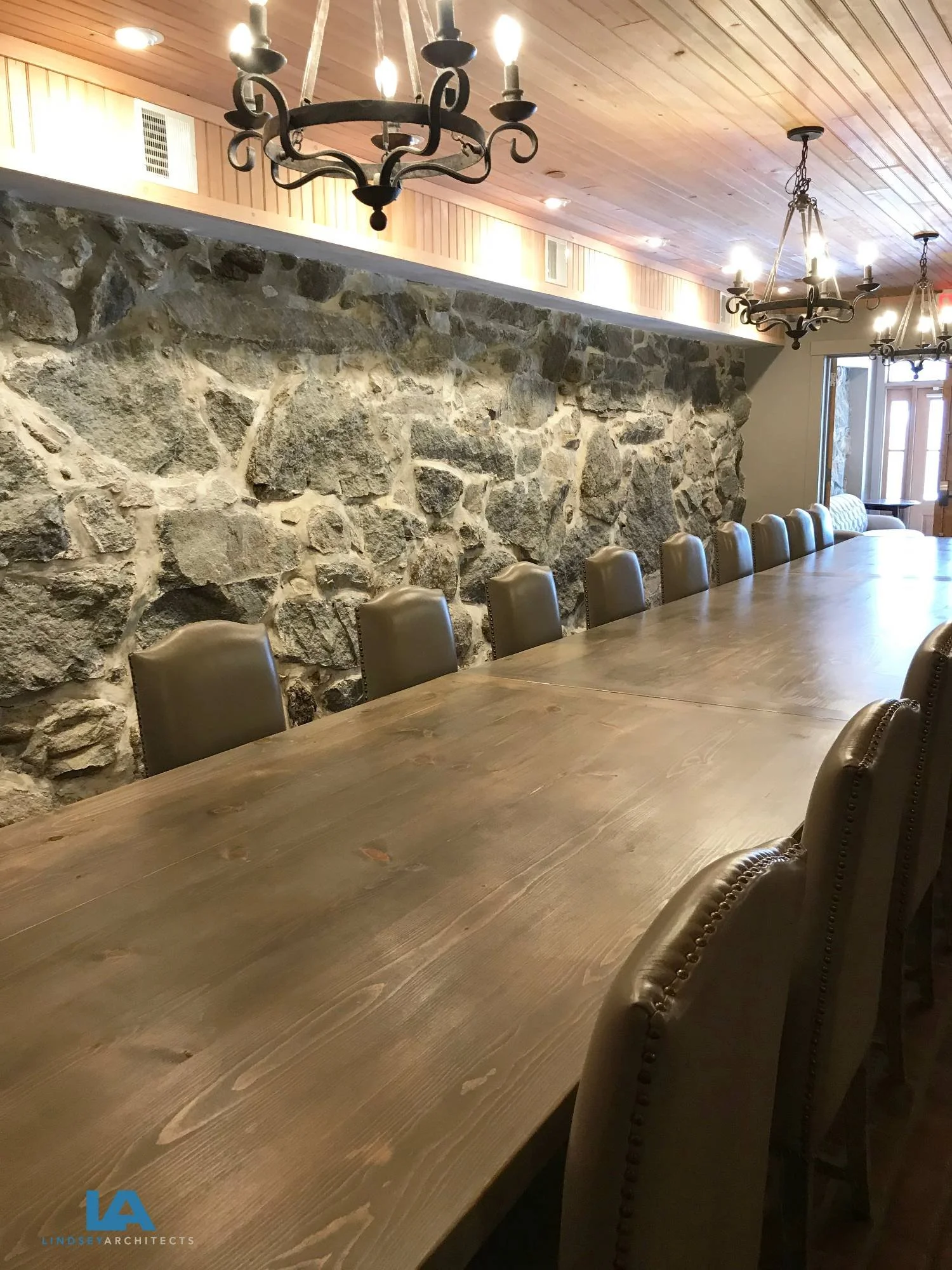 Elegant historic inn dining room featuring a dramatic original fieldstone accent wall, custom long farmhouse dining table with upholstered linen chairs, wrought iron chandeliers, white-painted wood-paneled ceiling, and warm ambient lighting — designe