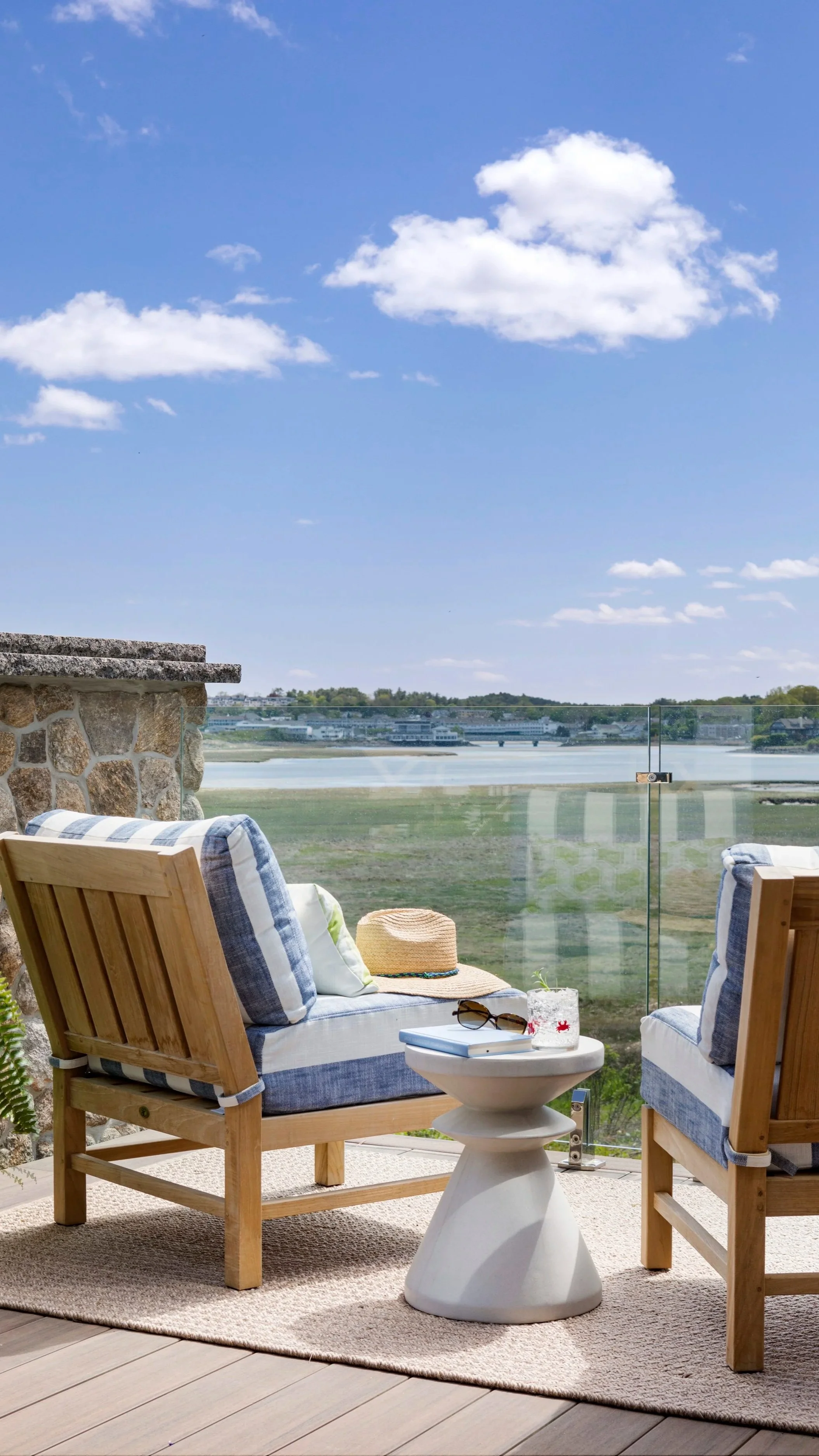 Teak outdoor lounge chair with blue cushions on a coastal deck overlooking an estuary and harbor, with a stone wall and blue sky — an outdoor living space designed by Lindsey Architects in Ogunquit, Maine.
