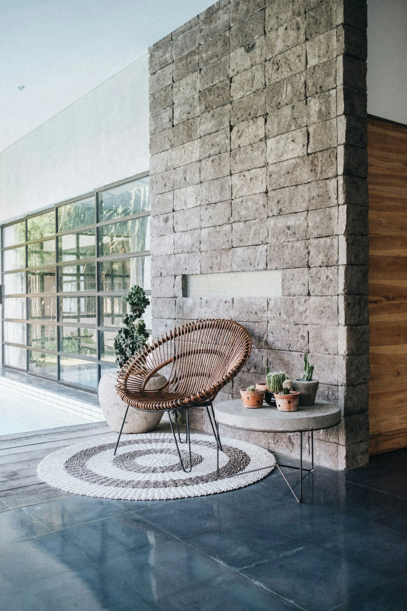 Indoor-outdoor terrace featuring a textured stone block wall, woven rattan lounge chair, round jute rug, and a steel-framed glass wall — illustrating the material continuity and seamless transitions Lindsey Architects designs into custom Maine homes.