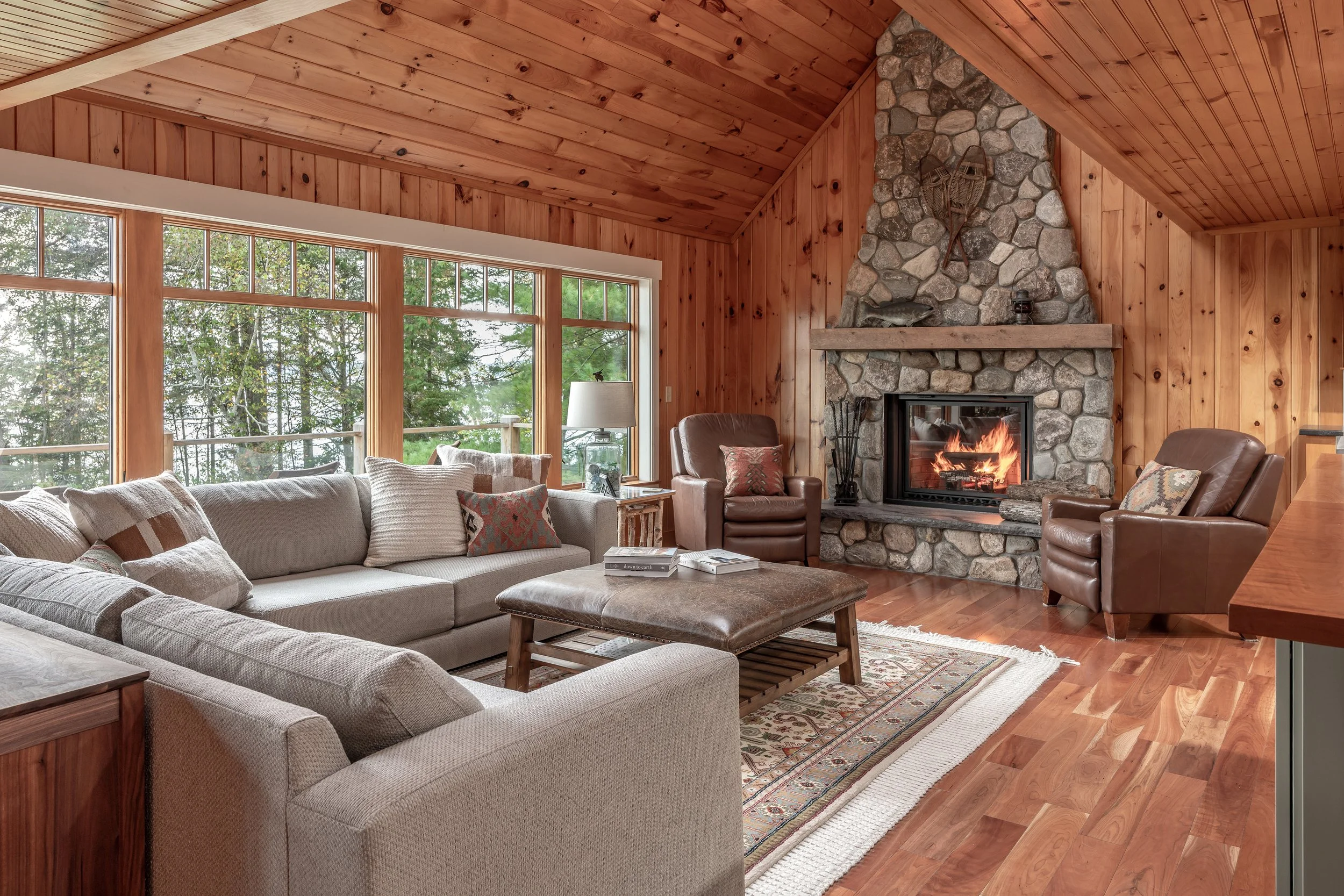 A warm and inviting great room designed by Lindsey Architects, featuring vaulted wood ceilings, expansive forest-facing windows, and a dramatic natural stone fireplace inspired by timeless Maine camp architecture. Rich wood textures, layered natural 