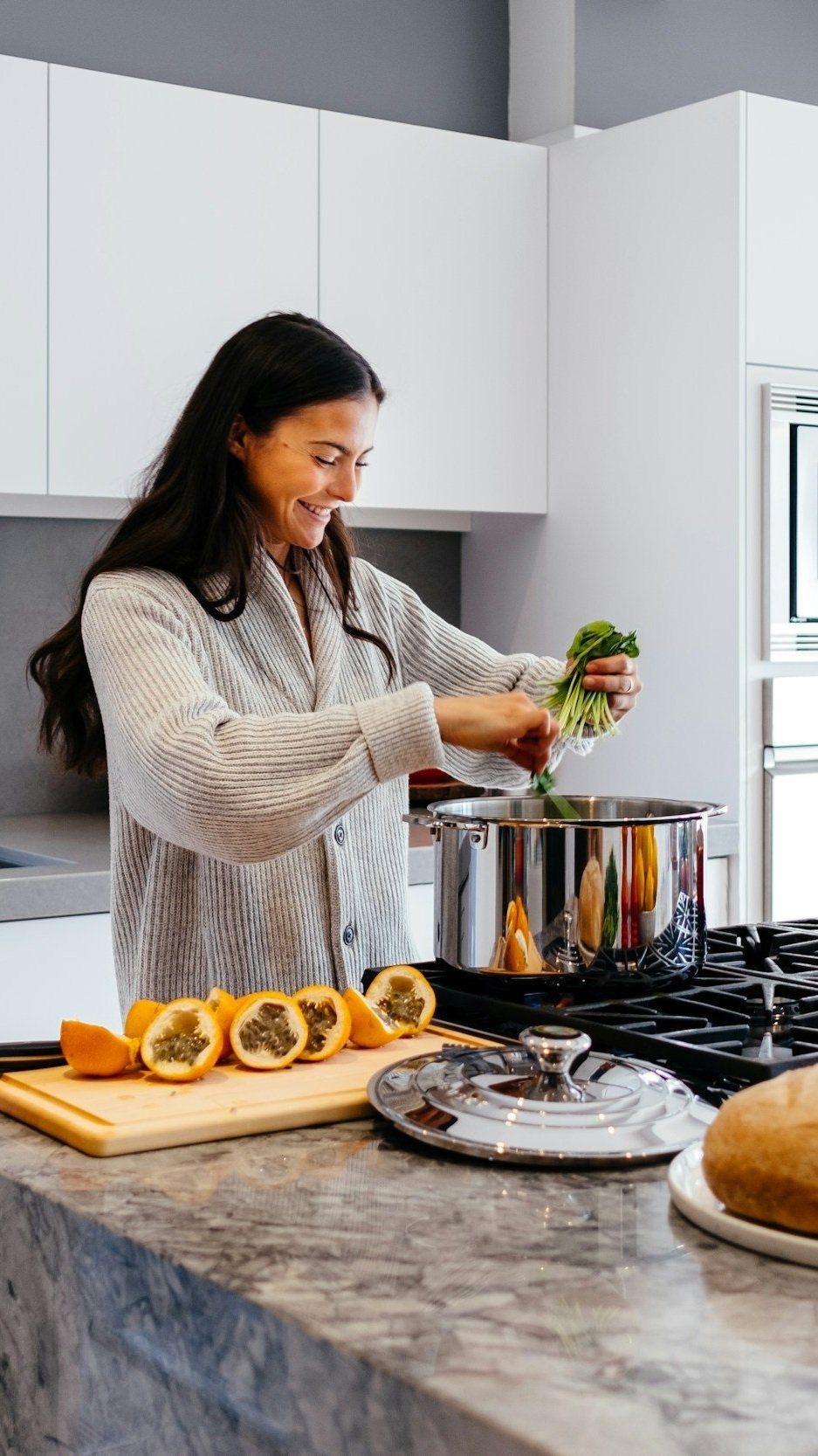 Woman preparing a meal at a gas range beside a marble-topped island with ample counter space — illustrating the generous, well-placed work surfaces Lindsey Architects designs into custom kitchens for coastal homes in Kennebunkport, Maine.
