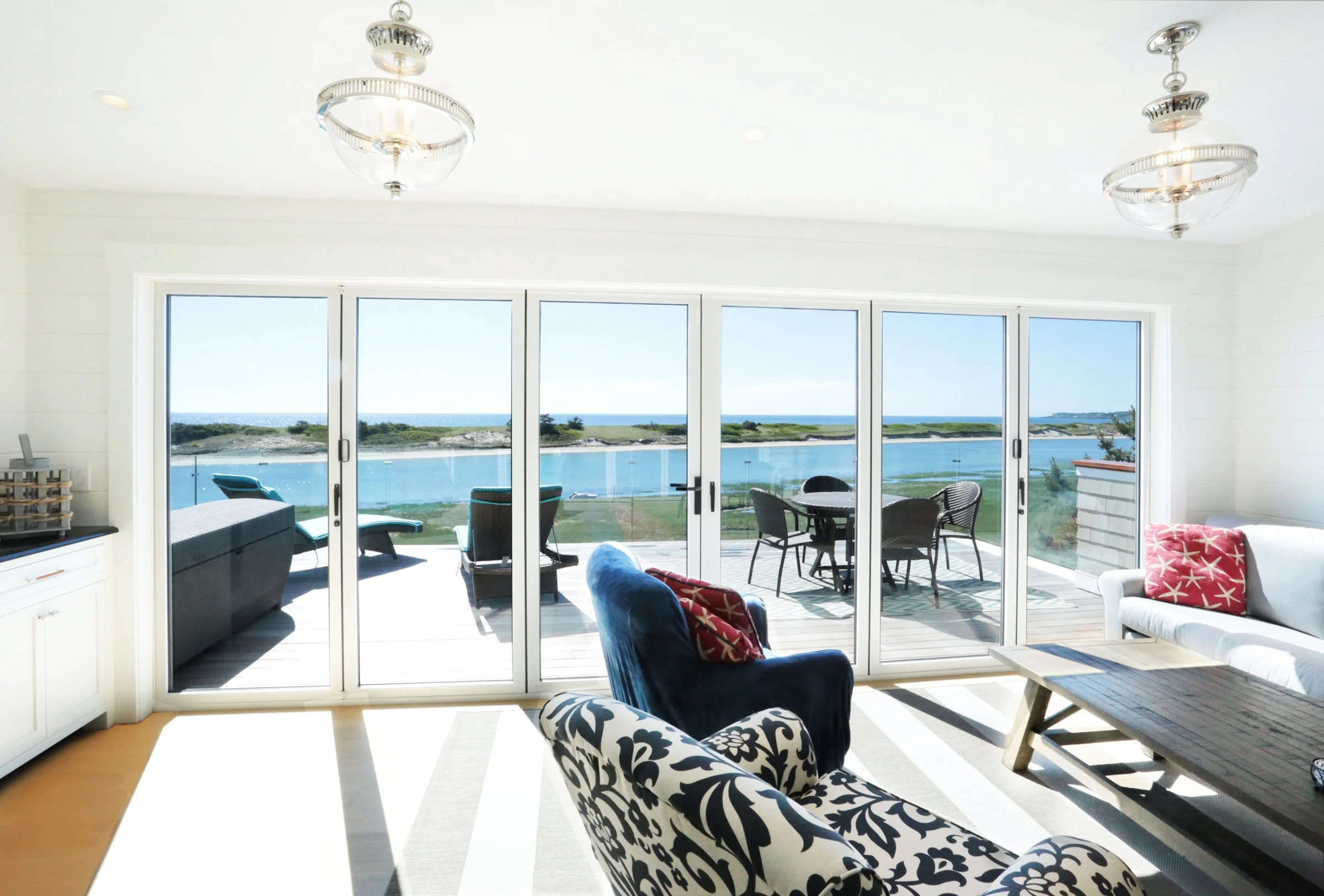 Luxury coastal living room interior featuring a wall of French doors opening to a private oceanfront deck with panoramic harbor views, crystal chandeliers, navy blue accent chair with patterned upholstery, white shiplap walls, and a built-in window s