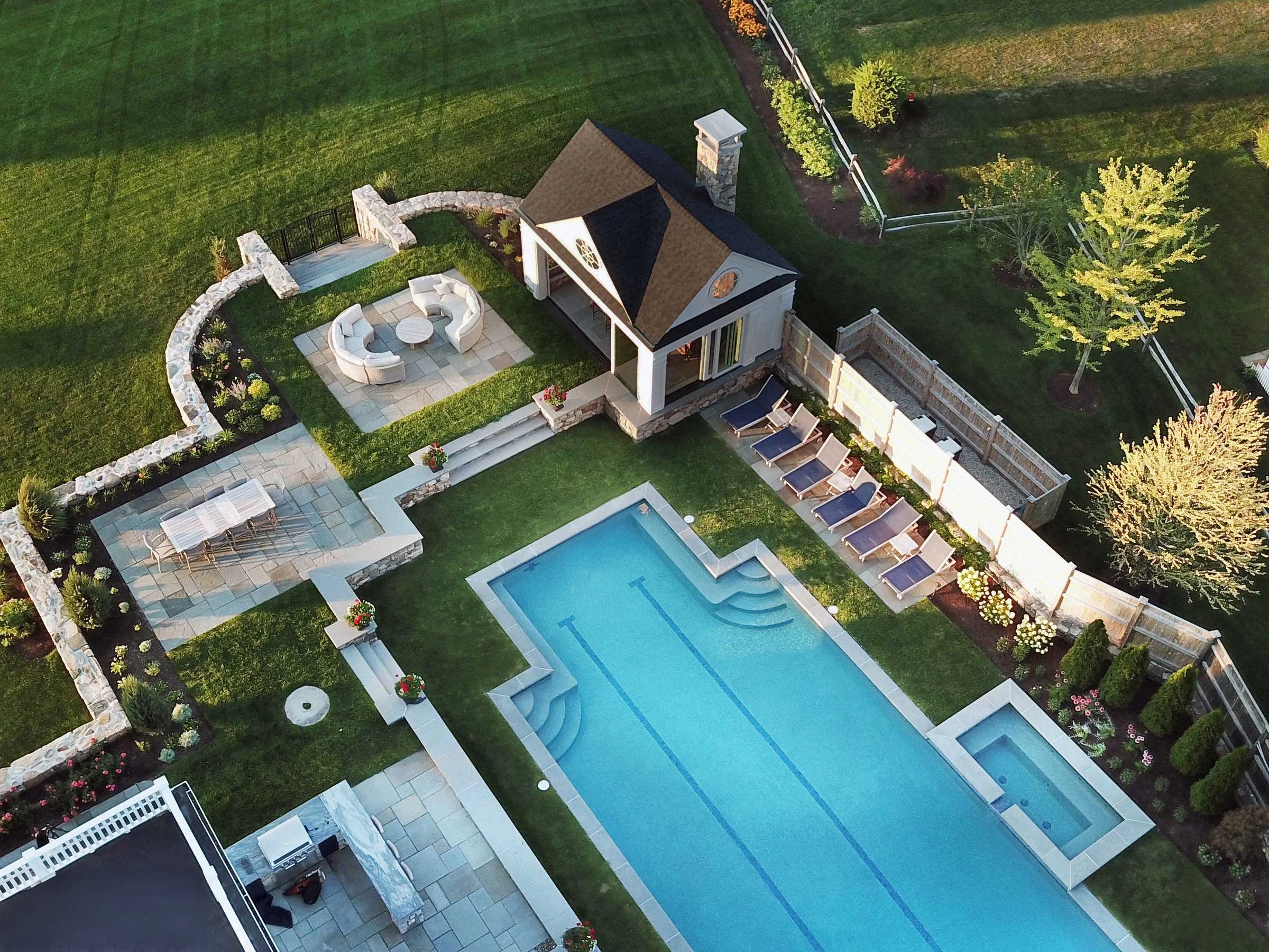 A backyard with a swimming pool, poolside lounge chairs, a patio with outdoor furniture, a small building with a bell, and well-maintained gardens and lawn.