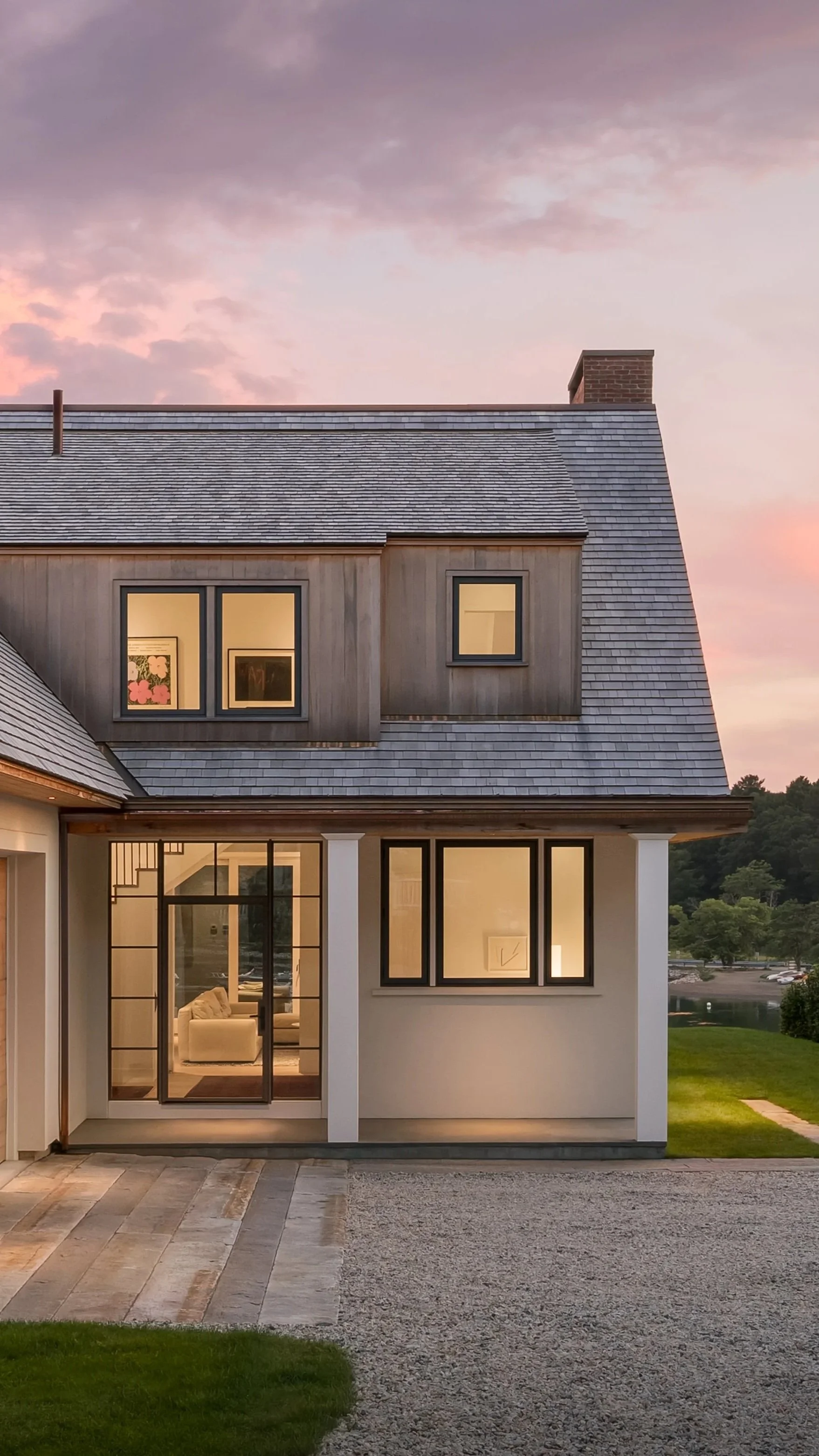 Cedar shake exterior of a custom home at dusk, with warm light glowing through steel-framed windows against a pink sky, designed by Lindsey Architects in York, Maine.