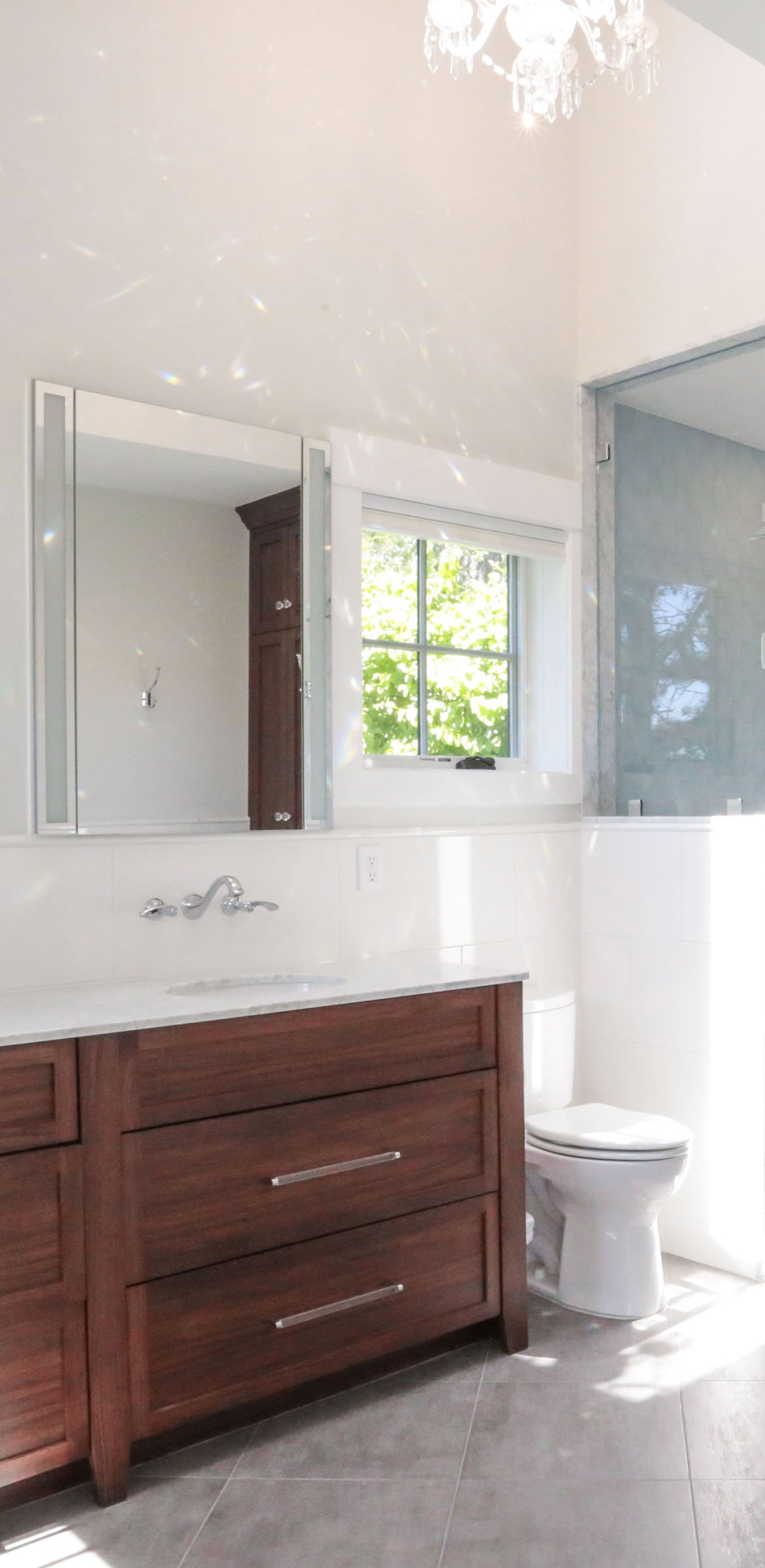 Luxury coastal primary bathroom featuring a custom dark walnut vanity with brushed nickel hardware, white quartz countertop, framed mirror, crystal chandelier, glass-enclosed shower, and a bright window framing natural greenery views. Designed by Lin