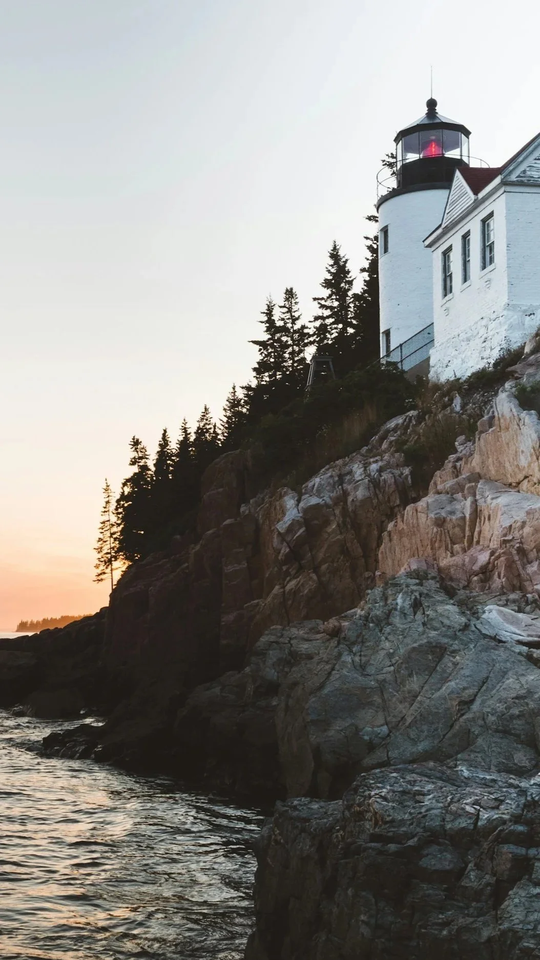 A white Maine lighthouse perched on dramatic granite ledge at dusk, surrounded by evergreens — illustrating the protected coastal environments governed by Maine's Shoreland Zoning Act, where Lindsey Architects designs custom homes.