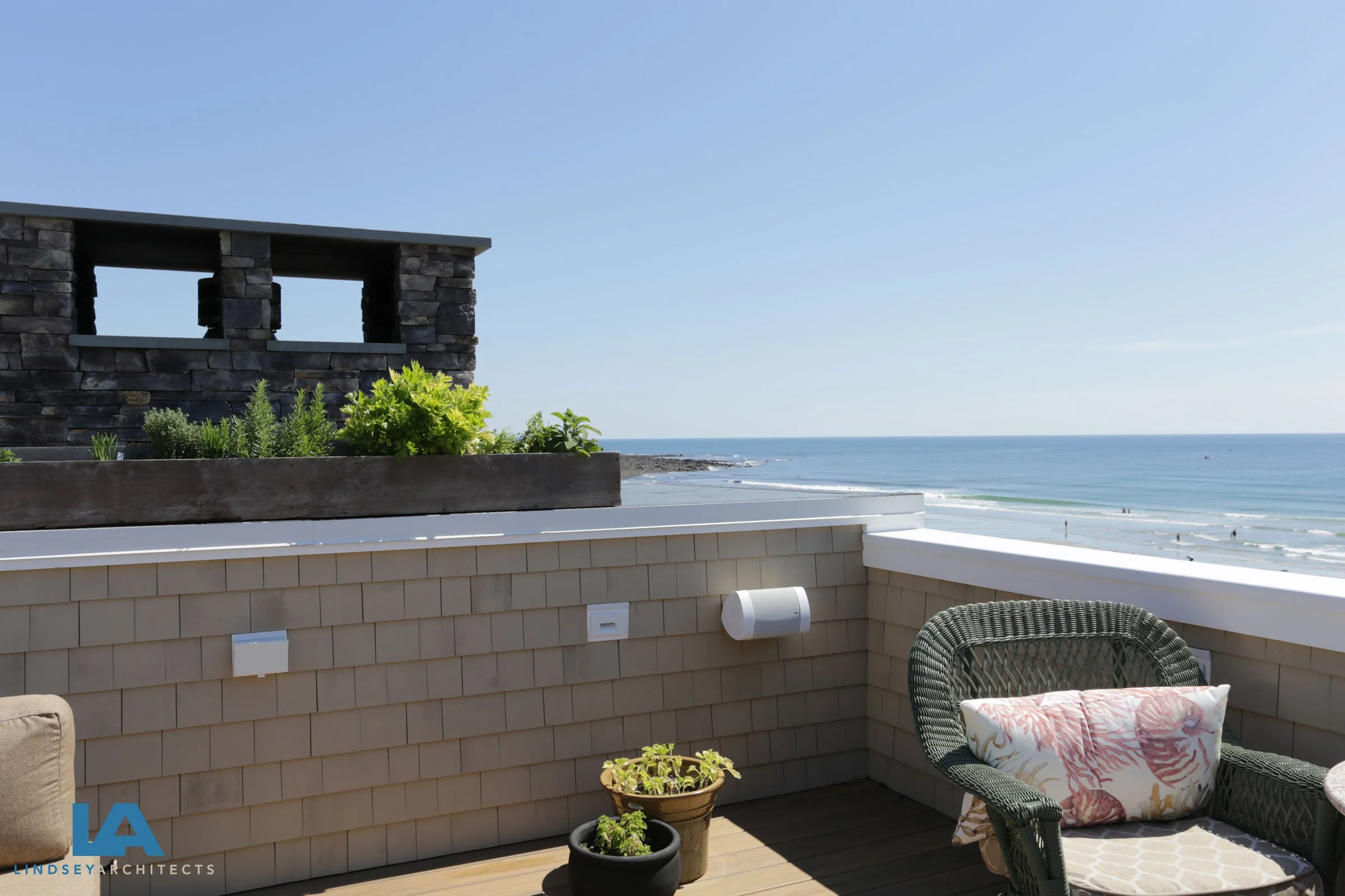 Private rooftop deck with panoramic Atlantic Ocean Views featuring cedar shingle siding, modern stone chimney with built in planter woven wicker seating, and potted greenery, custom luxury coastal home by Lindsey Architects, serving Kennebunkport