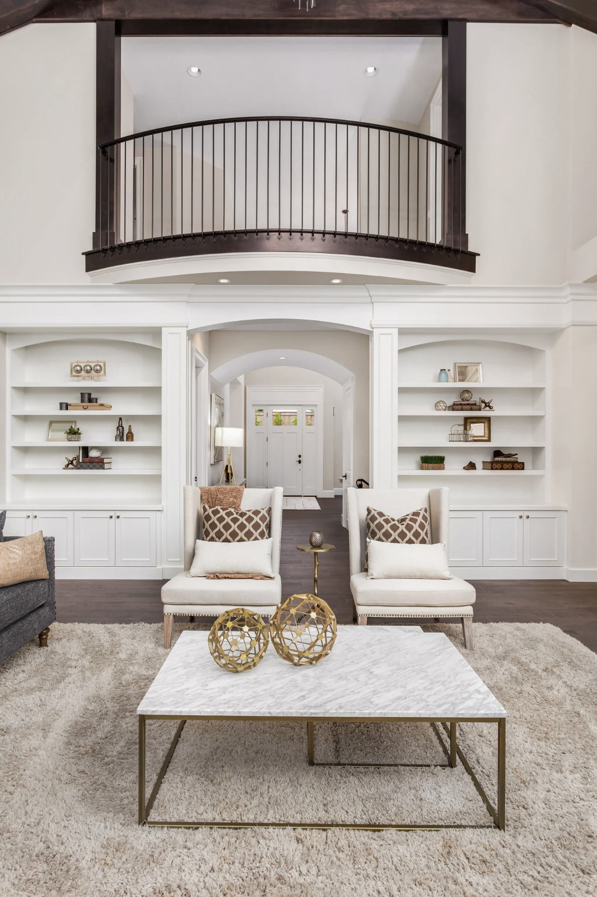 Custom home great room by Lindsey Architects featuring floor-to-ceiling built-in bookshelves, a curved wrought-iron balcony, vaulted ceiling with exposed beams, and arched hallway — showcasing thoughtful space planning in a coastal Maine residence.