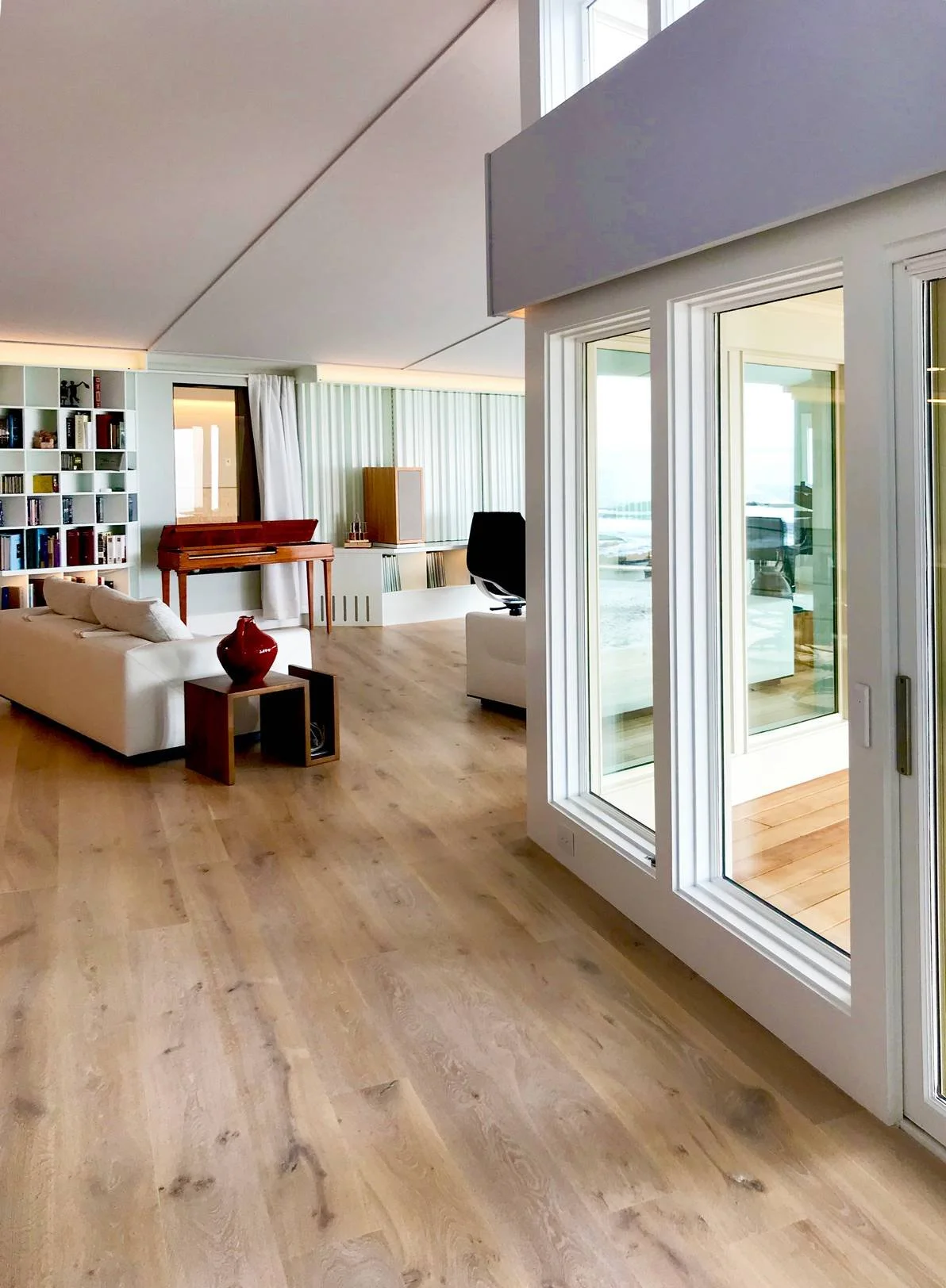 Open-concept modern coastal living space featuring wide-plank white oak flooring, a vaulted ceiling, floor-to-ceiling glass doors opening to an oceanfront deck, white modern sectional seating, custom built-in shelving, and abundant natural light — cu