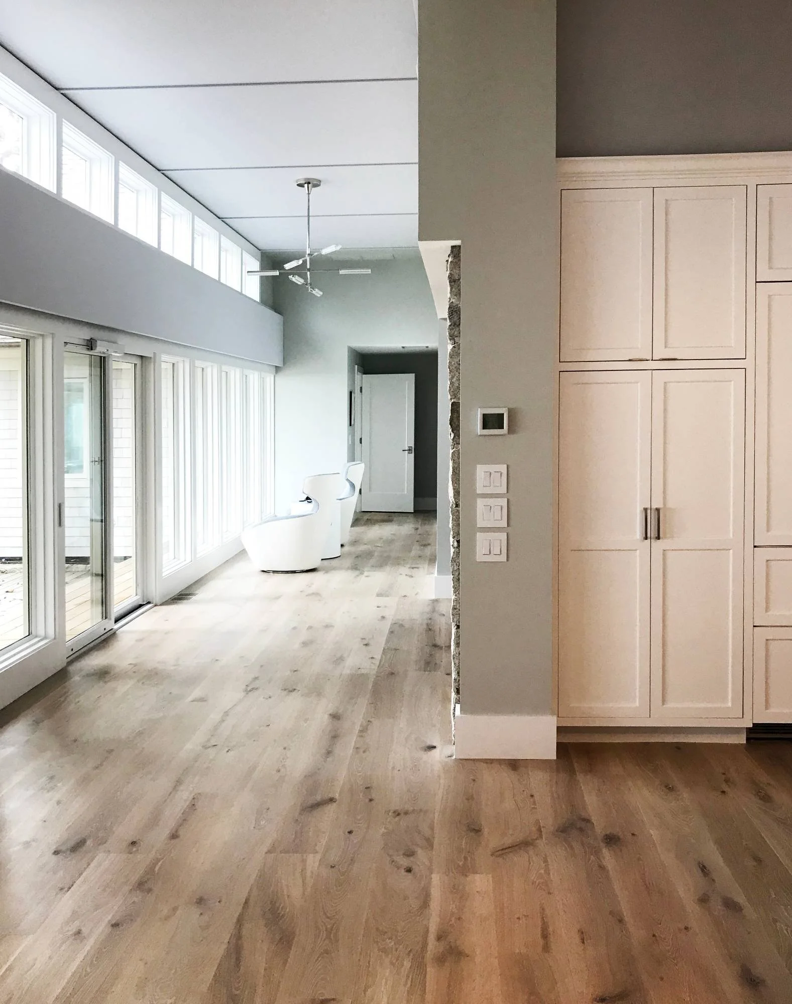 Bright open-concept coastal interior featuring wide-plank white oak flooring, a soaring vaulted ceiling, custom built-in white cabinetry, and a wall of windows leading to a sun-filled living area with ocean views — custom luxury coastal home by Linds