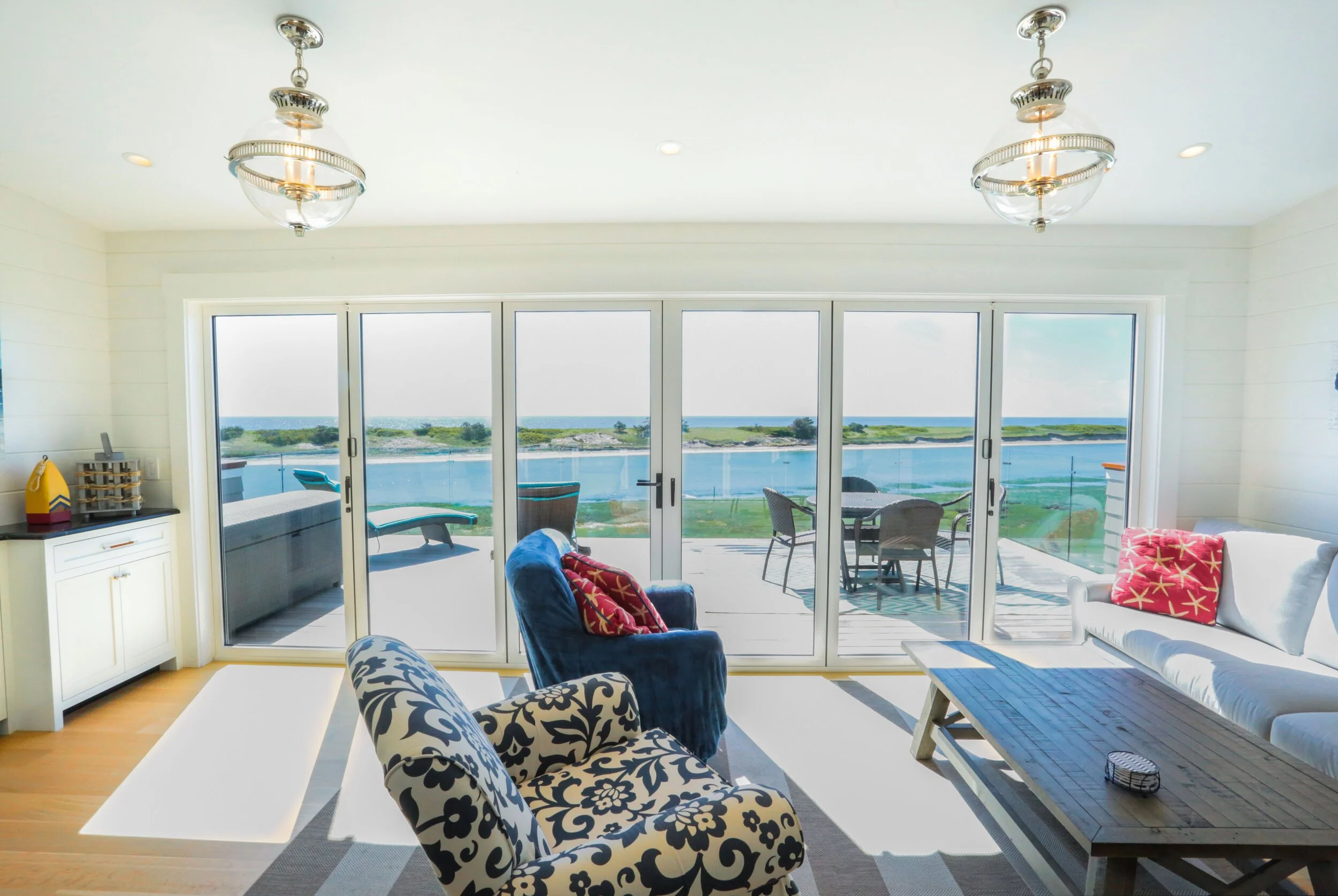 Living room with large sliding glass doors opening to a balcony overlooking water and greenery. The room has patterned armchairs, a white couch with red pillows, a blue coffee table, and a small cabinet with a yellow bag on top. Two modern ceiling lights hang from the ceiling.