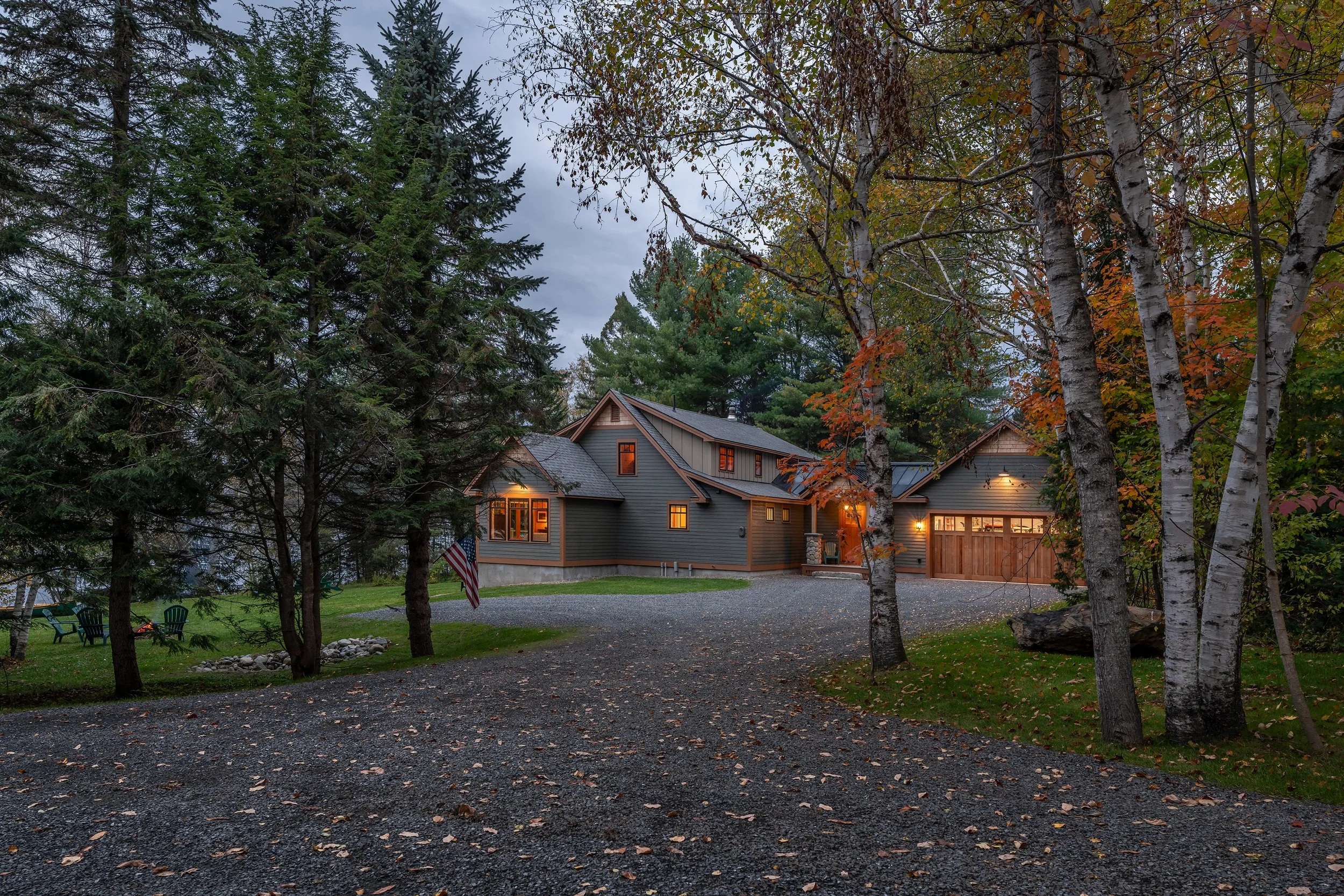 A custom woodland residence designed by Lindsey Architects, thoughtfully integrated into the natural Maine landscape with warm exterior lighting, timeless architectural forms, and carefully crafted materials. Surrounded by mature trees and approached