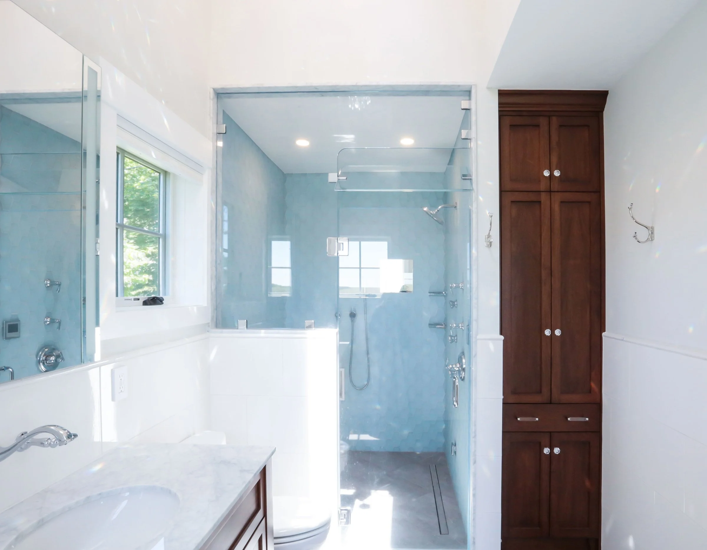 A refined contemporary bathroom designed by Lindsey Architects, featuring custom walnut storage cabinetry, a frameless glass shower enclosure, and soft natural light inspired by elegant coastal living in Maine and New Hampshire. The spa-inspired layo