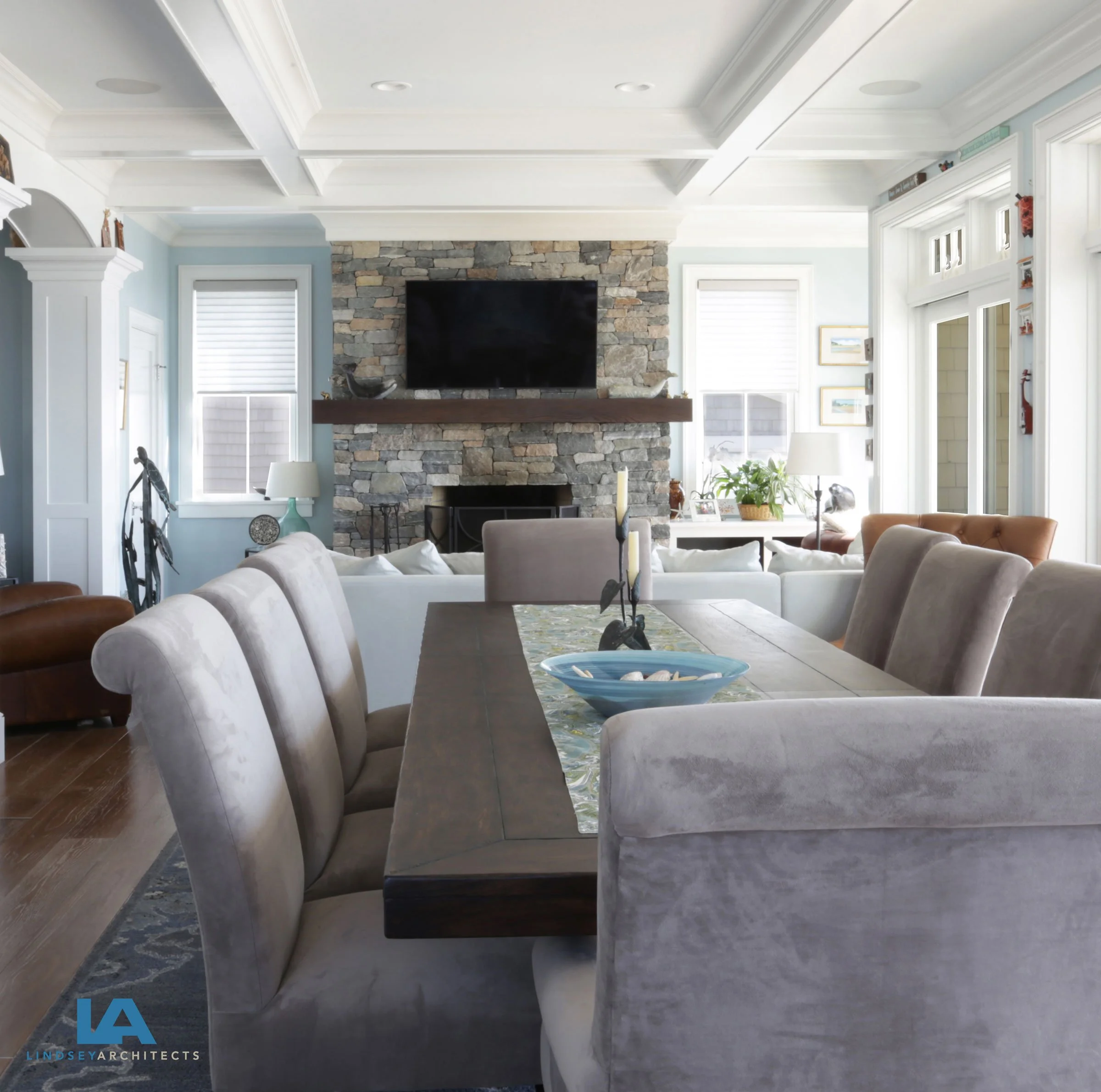 Coastal luxury great room featuring an open-concept dining and living area, long rustic farmhouse table with gray velvet upholstered chairs, stacked stone fireplace with mounted television, coffered ceiling with white beam detailing, soft blue accent