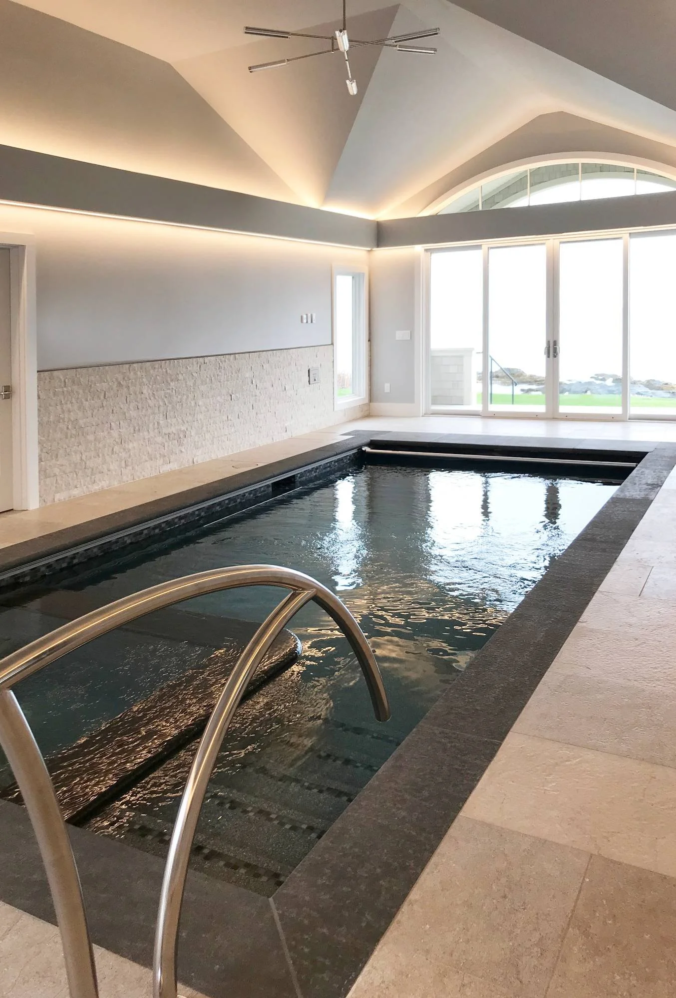 rivate indoor lap pool featuring a travertine tile deck and surround, vaulted ceiling with recessed cove lighting, stainless steel handrails, and oversized windows framing panoramic ocean views — designed for year-round wellness and seaside living. C