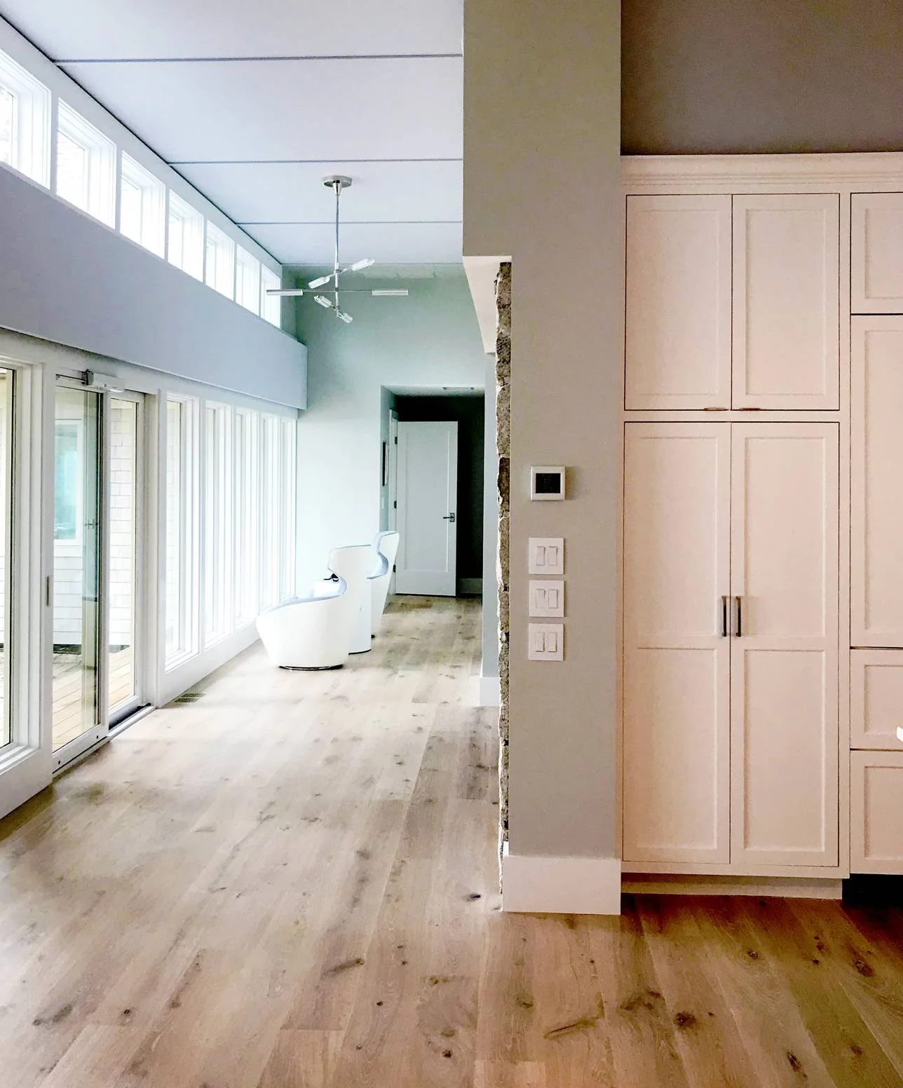 Bright open-concept coastal interior featuring wide-plank white oak flooring, a soaring vaulted ceiling with clerestory windows, floor-to-ceiling glass doors opening to an oceanfront deck, custom built-in white cabinetry, and a sun-filled adjoining l