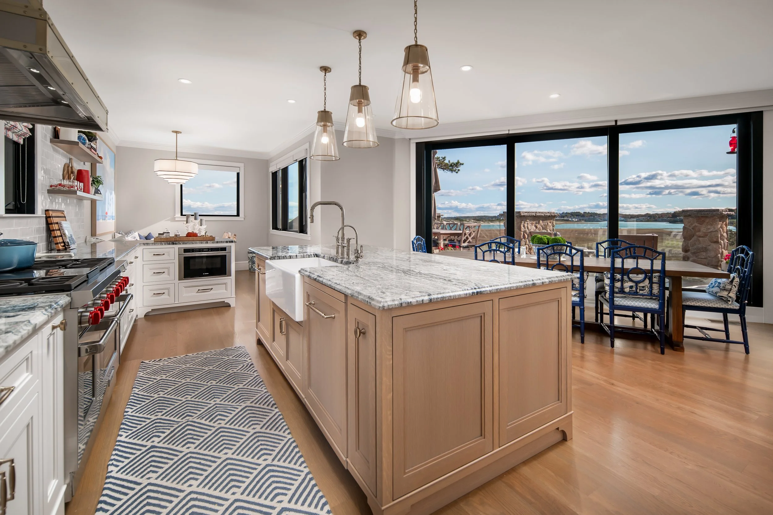 Bright kitchen with white cabinets, a large island with a marble top, blue chairs around a dining table, large windows showing a view of water and sky, pendant lighting over the island, and open shelving on the wall.
