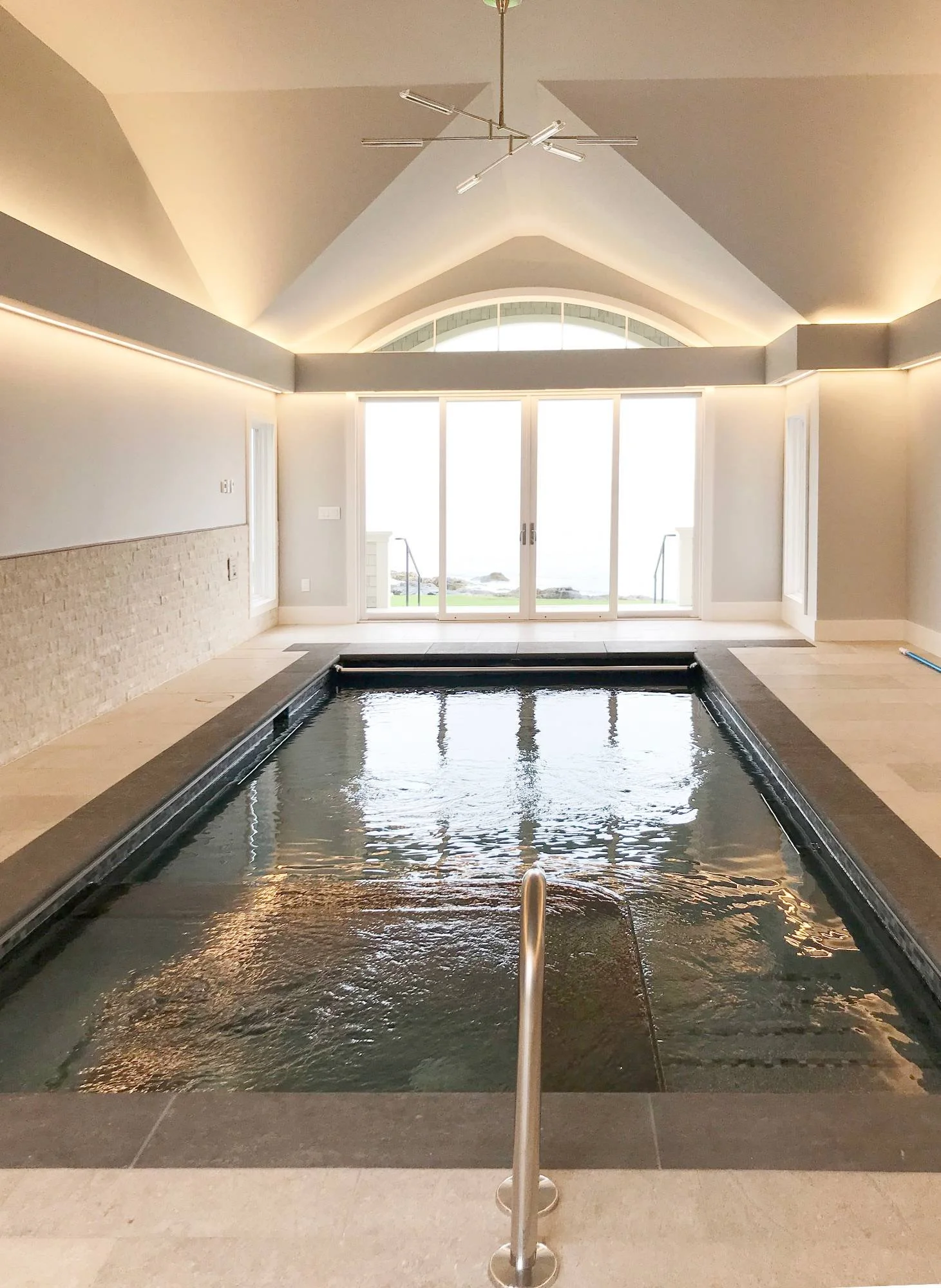 Private indoor lap pool featuring a vaulted ceiling with recessed cove lighting, travertine tile surround, stainless steel handrail, and French doors framing ocean views — designed for year-round wellness in a custom luxury coastal home by Lindsey Ar