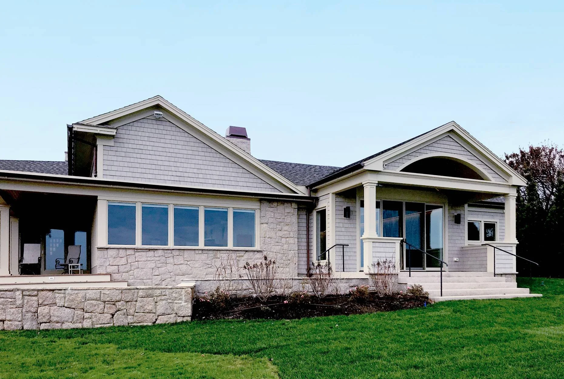 Custom luxury single-story coastal home exterior featuring gray cedar shingle siding, a natural stone foundation, an arched covered entry portico, gabled rooflines, expansive picture windows for natural light, a covered side porch with white columns,