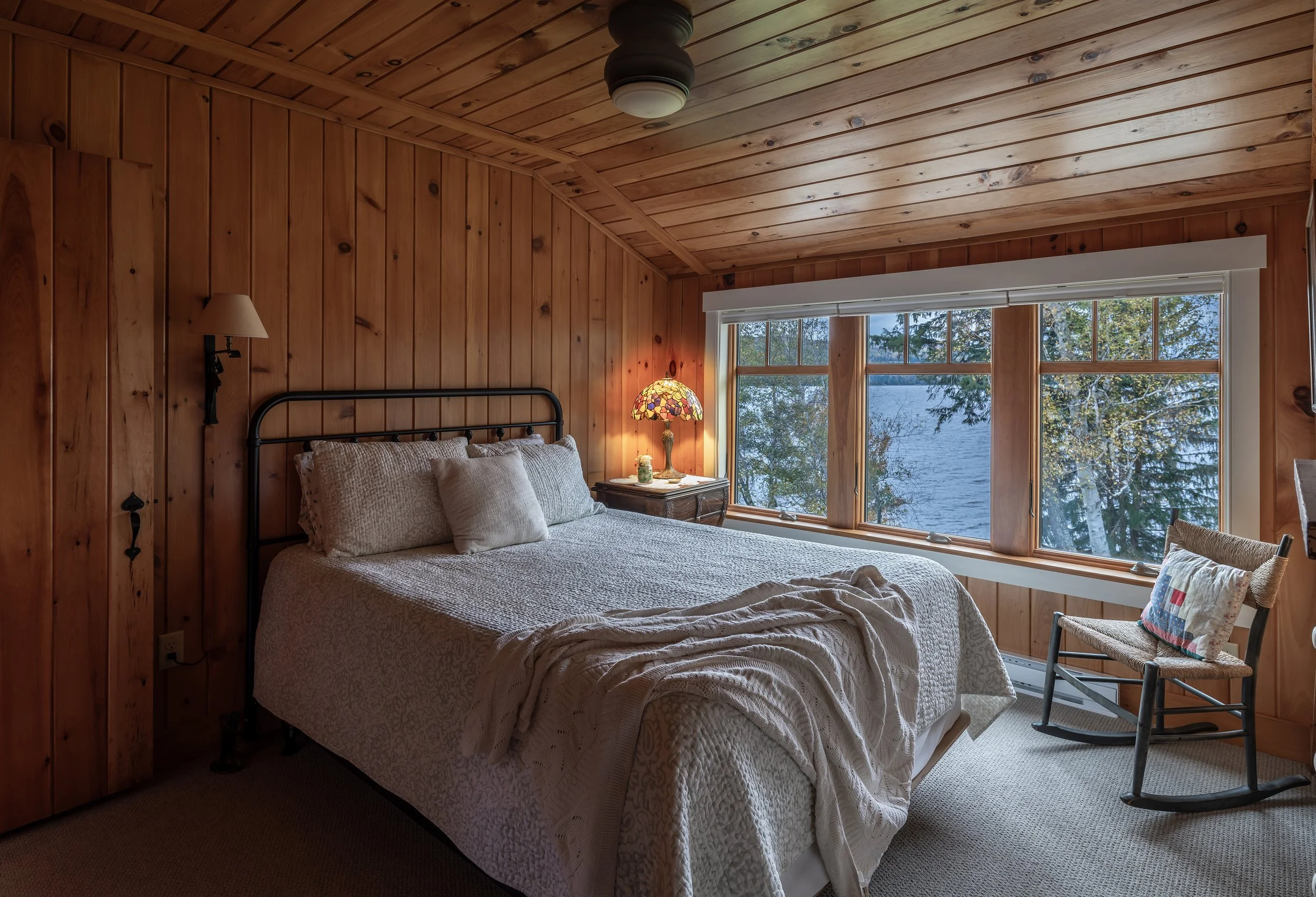 A warm and intimate lakeside bedroom designed by Lindsey Architects, featuring natural wood walls and ceilings, expansive waterfront windows, and timeless cabin-inspired detailing. Soft layered textiles, warm ambient lighting, and carefully framed vi
