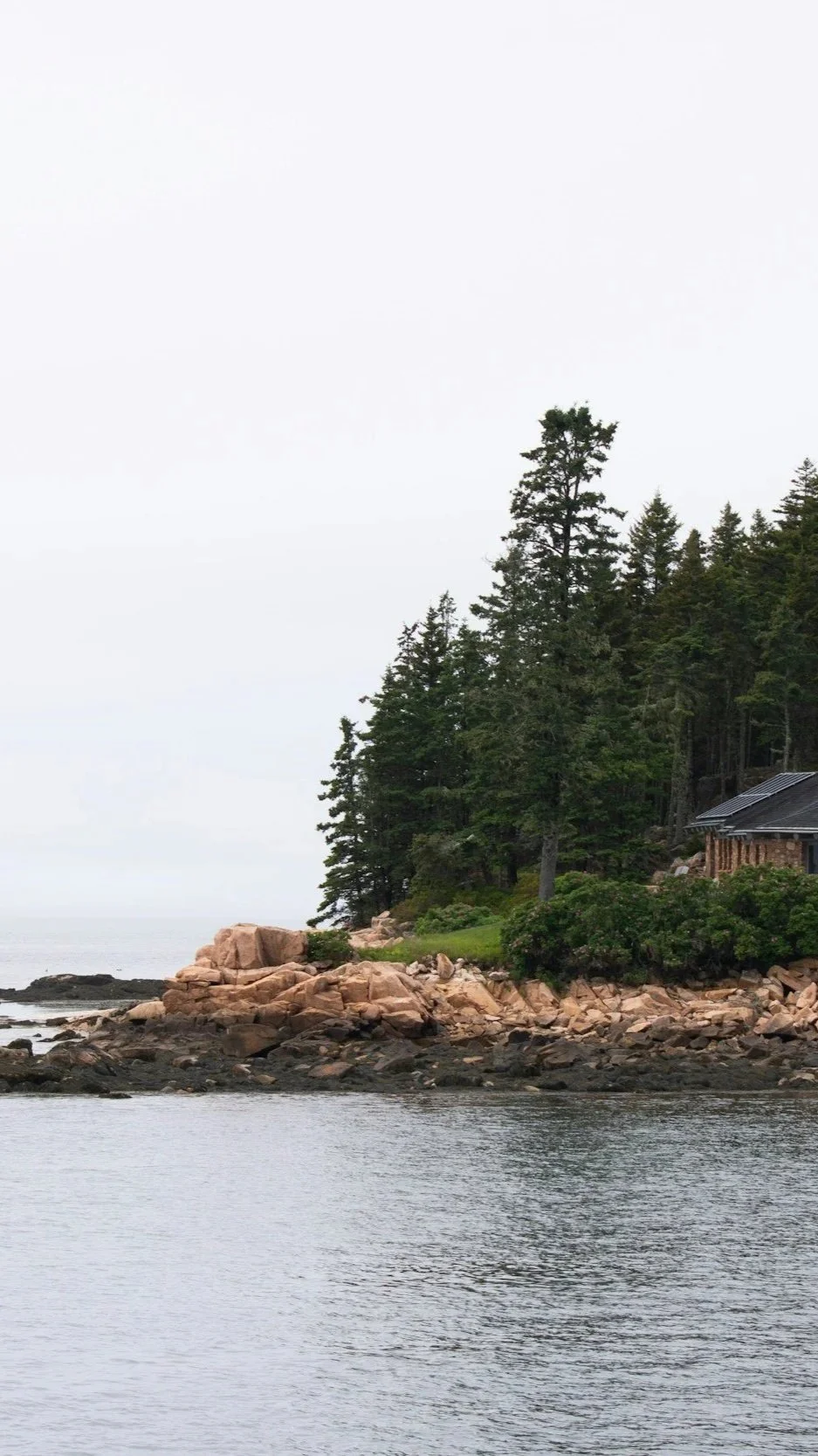 A coastal Maine home nestled among granite ledge and evergreen trees on a rocky waterfront point, viewed from the water — illustrating the site-specific, resilient coastal architecture Lindsey Architects designs in Kittery Point, Maine.