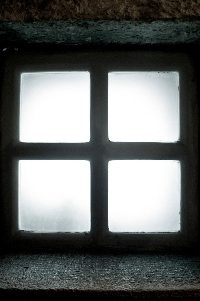 Small four-pane window in a dark stone wall letting in limited light — illustrating the common design mistake of poor window positioning that Lindsey Architects avoids through site-oriented window placement in custom Maine homes.